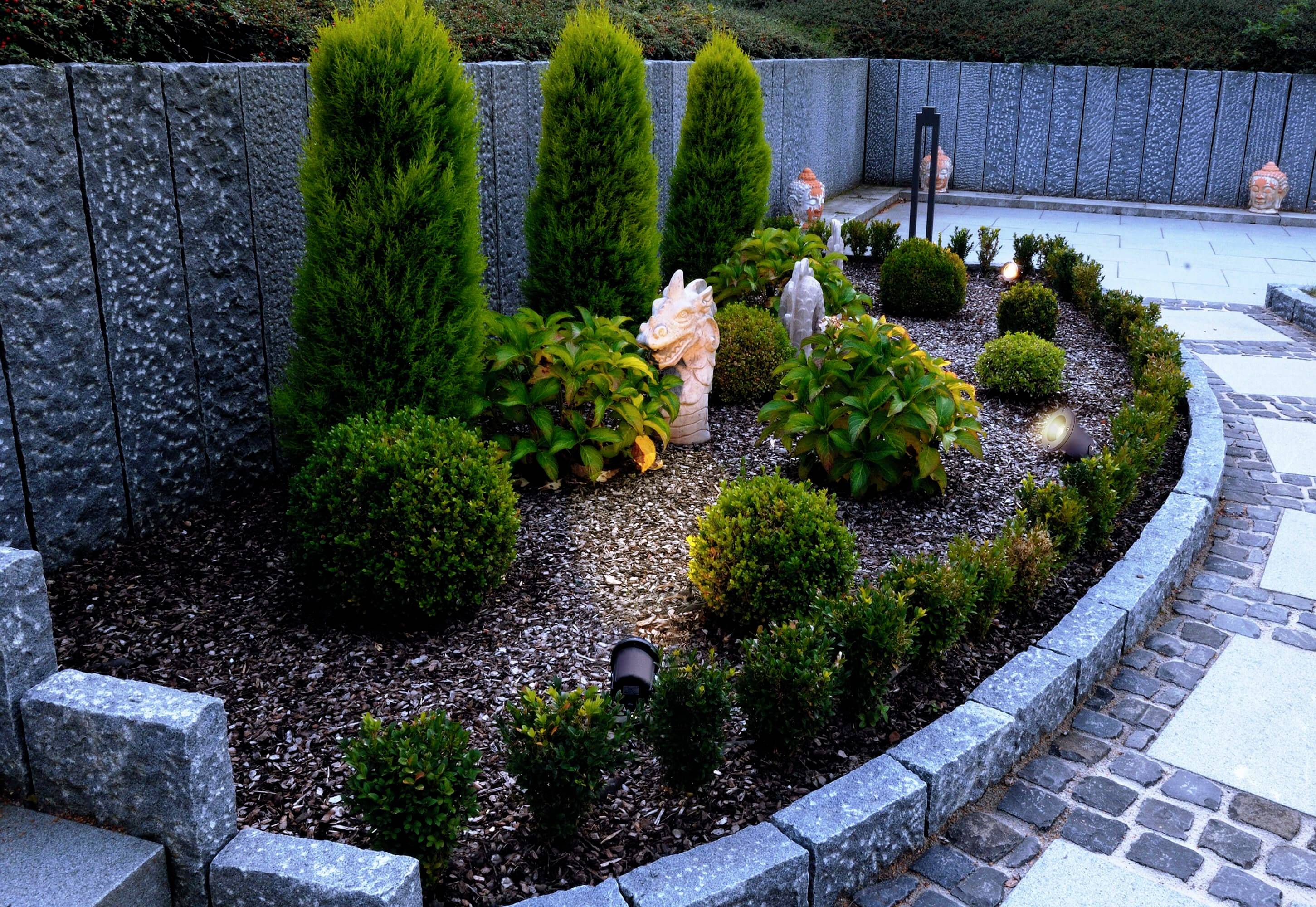 A well-maintained bed with evergreen shrubs and trees, surrounded by stone walls and paving, decorated with a small statue.