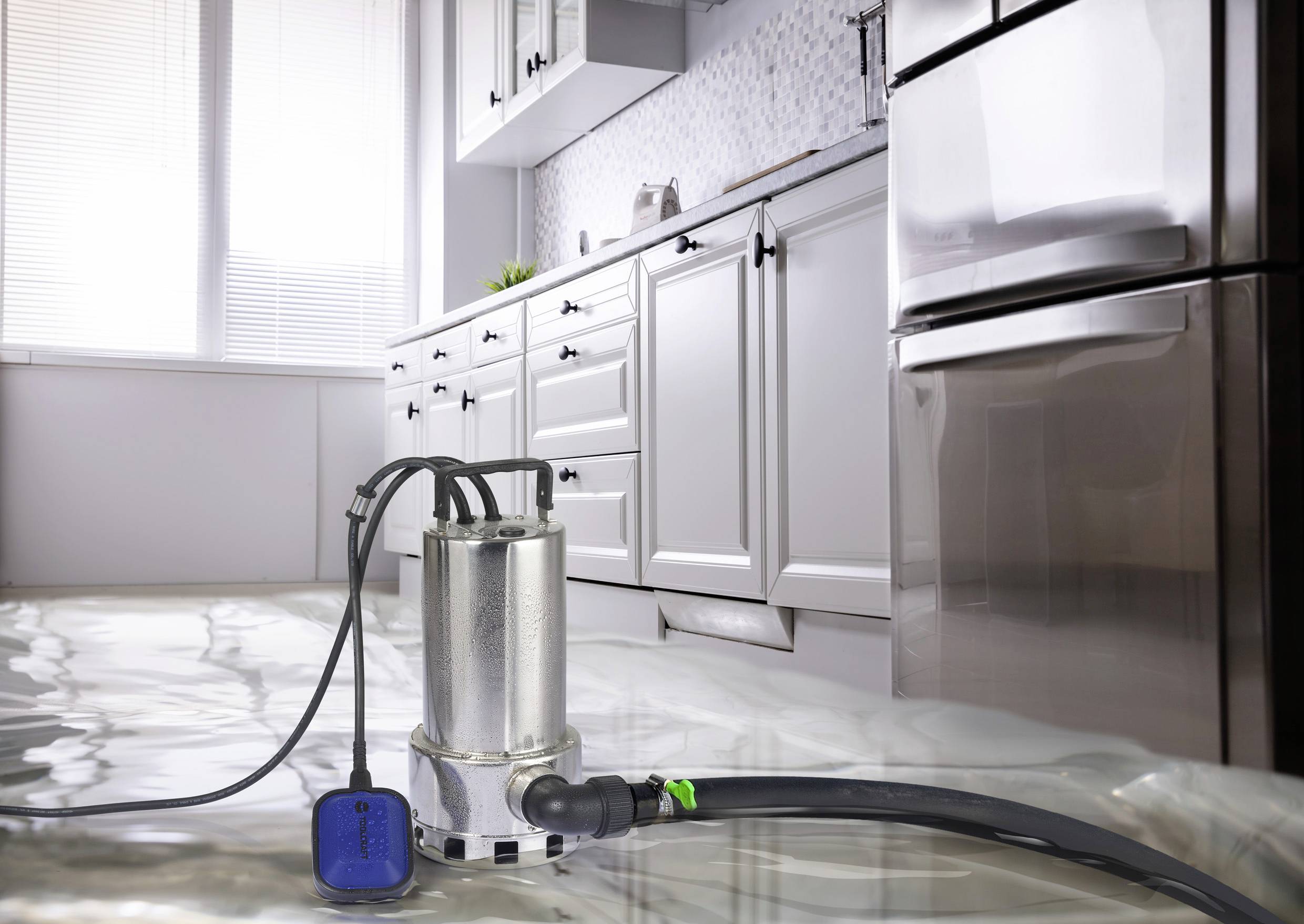A water pump is pumping water out of a flooded kitchen, with water standing on the floor. Cupboards and appliances are visible in the background.