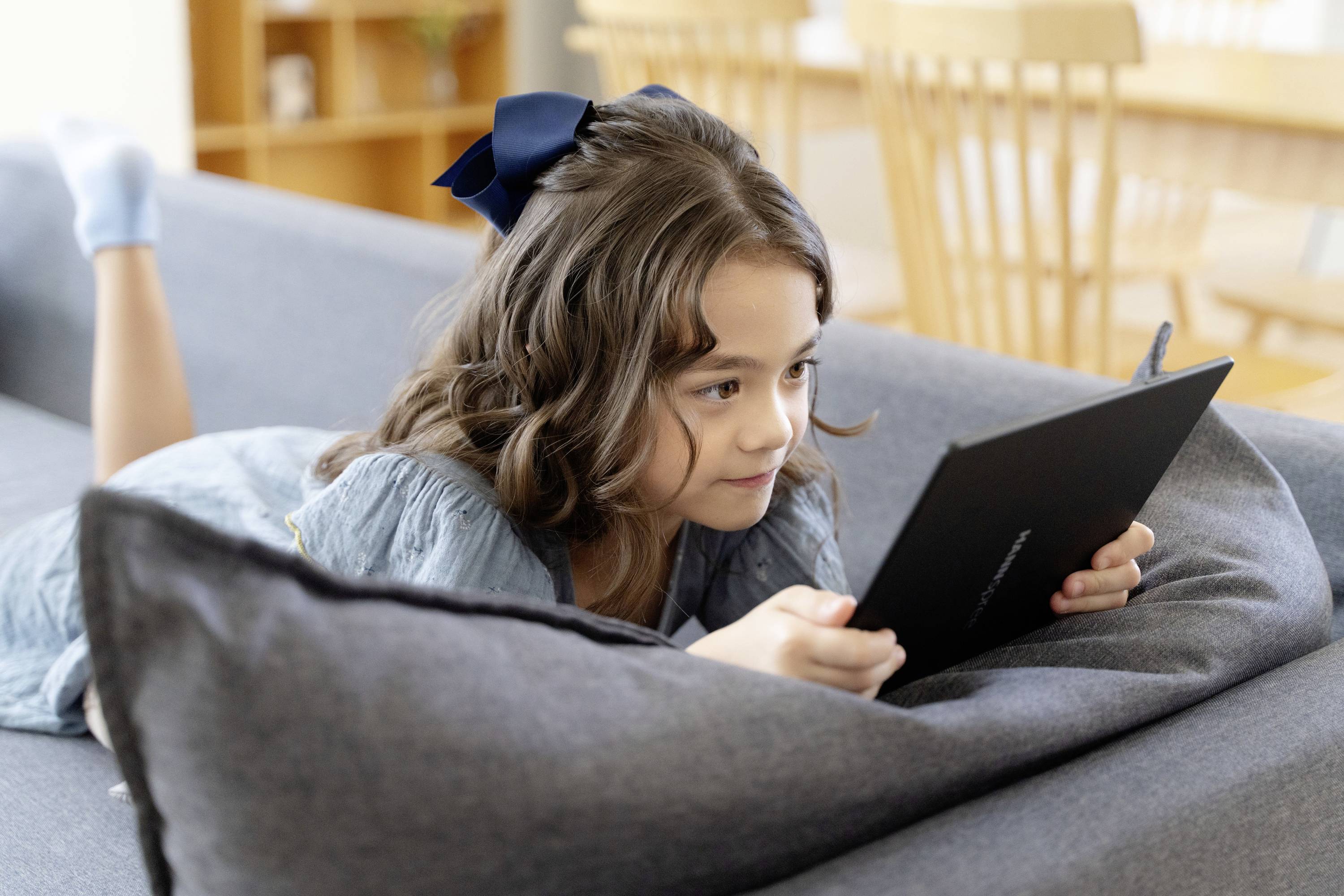 A girl is lying on a sofa and reading or playing on a tablet. She has brown hair and is wearing a hair bow.