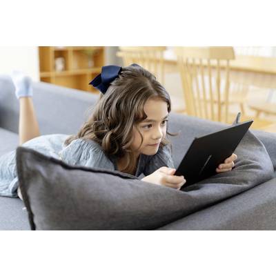 A girl is lying on a sofa and reading or playing on a tablet. She has brown hair and is wearing a hair bow.