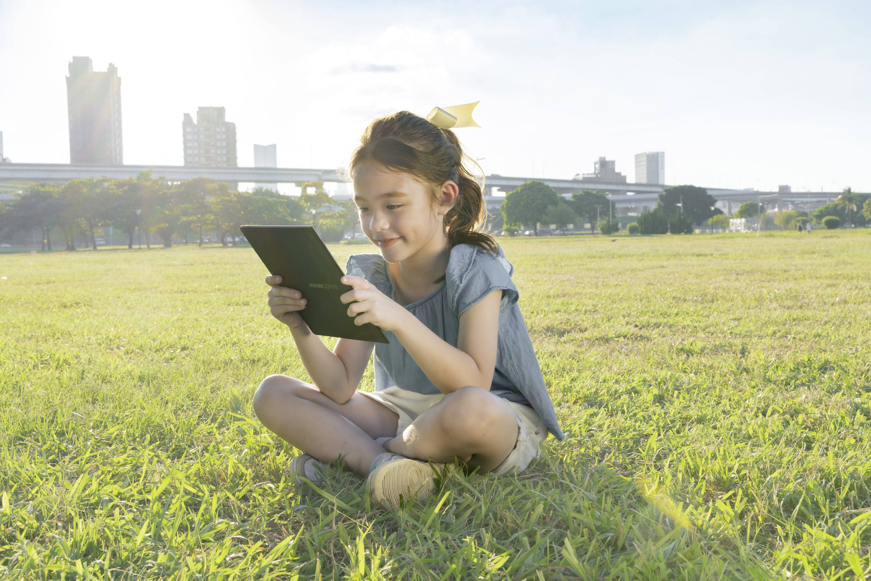 A girl is sitting on a grassy patch and looking at a tablet. Office buildings and a sunny sky can be seen in the background.