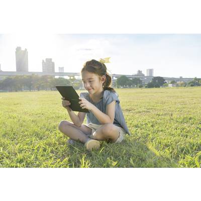 A girl is sitting on a grassy patch and looking at a tablet. Office buildings and a sunny sky can be seen in the background.