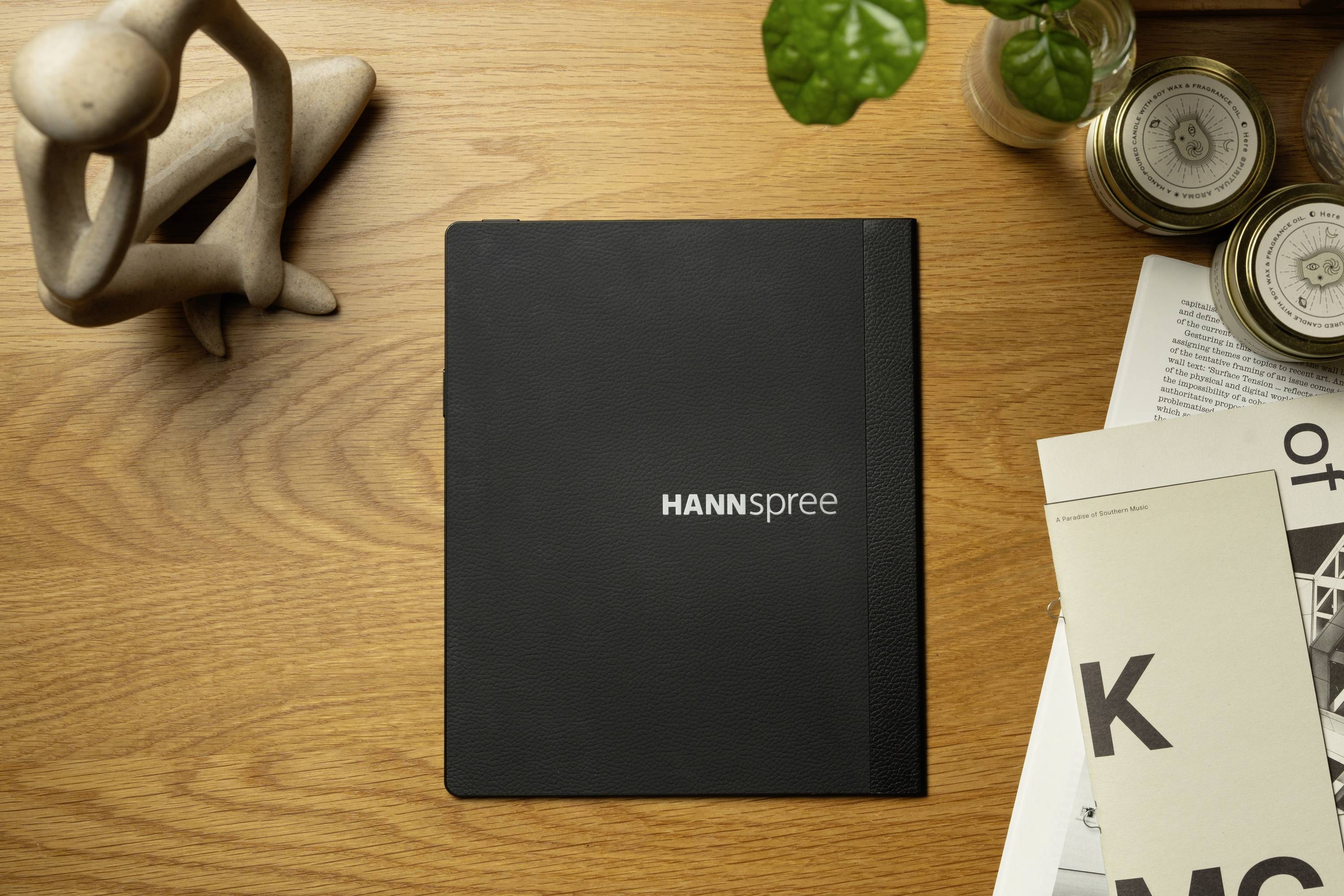 Black notebook with 'HANNSpree' written on it lies on a wooden table, surrounded by magazines, a small wooden model and a plant.