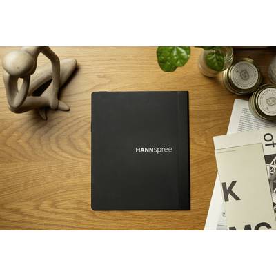 Black notebook with 'HANNSpree' written on it lies on a wooden table, surrounded by magazines, a small wooden model and a plant.