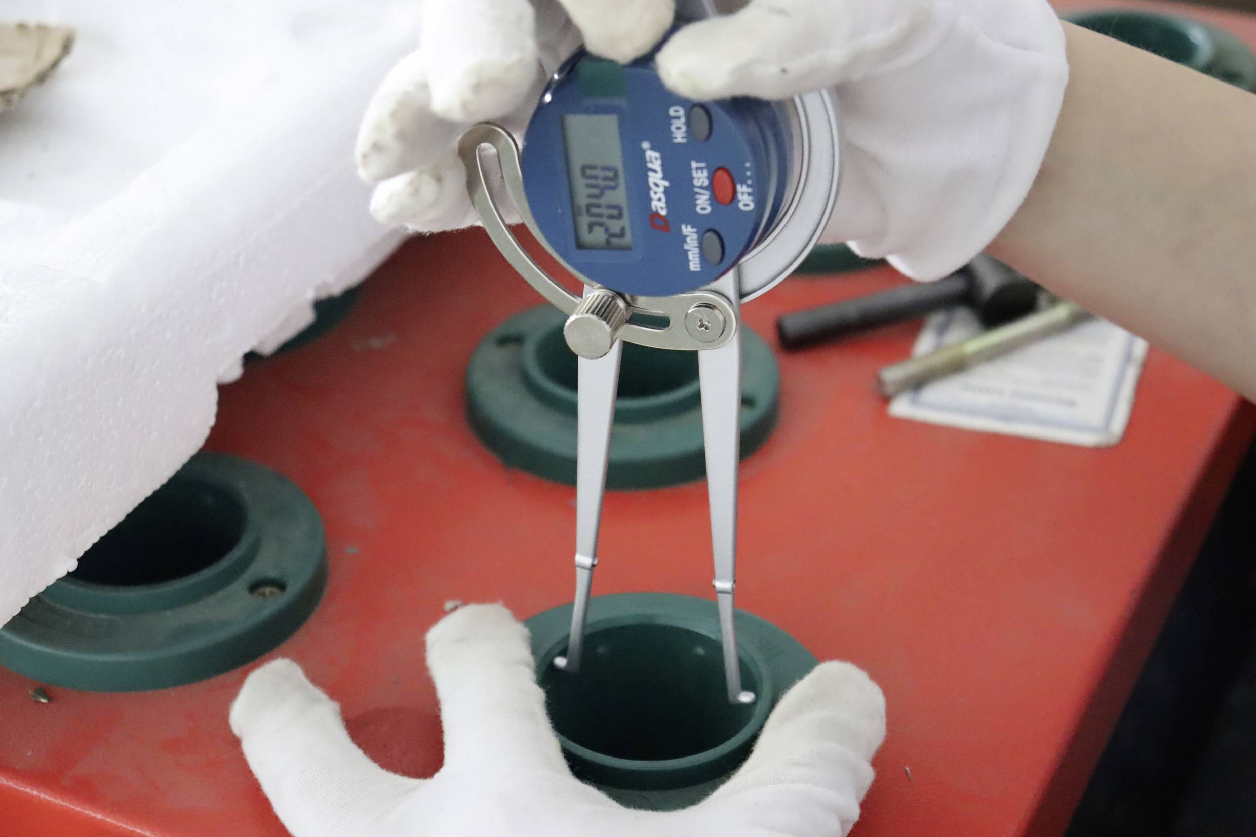 A person is measuring the internal diameter of a green cylinder on a red surface using a digital measuring instrument, wearing gloves.