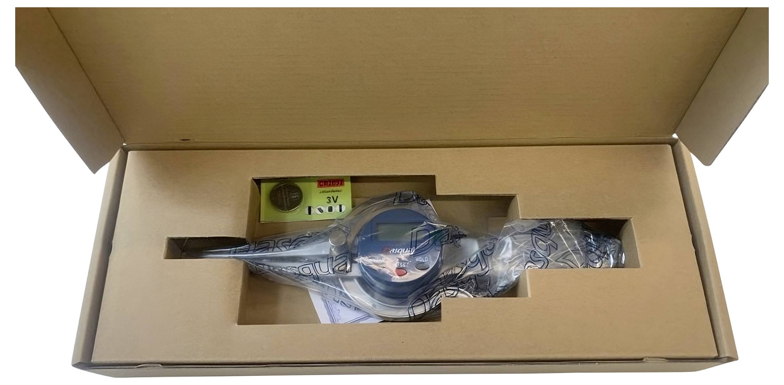 A water meter in a cardboard box, partially covered in plastic. Battery and coin are visible next to the meter.