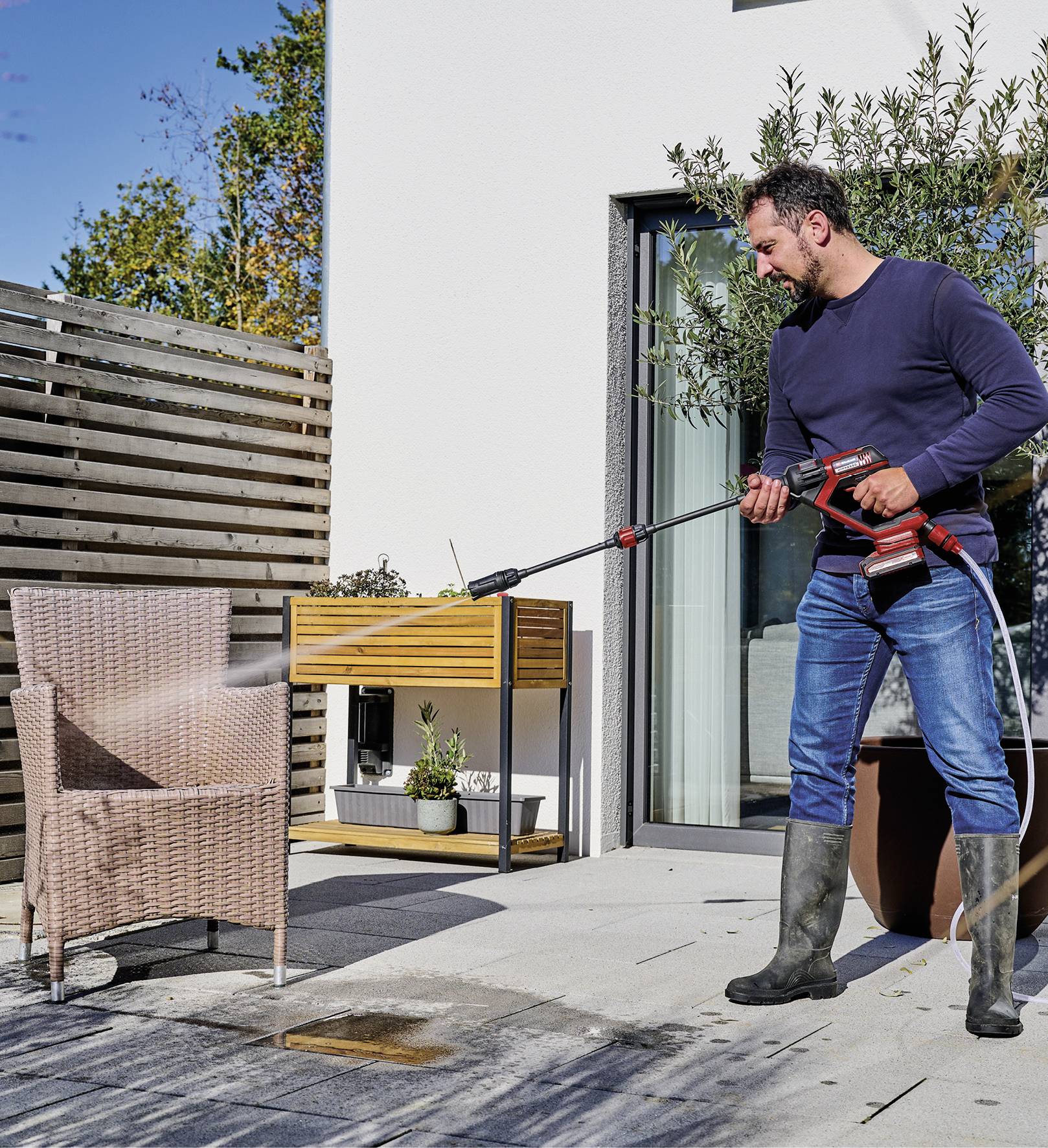 A man is cleaning a woven chair with a pressure washer on a patio. Plants and furniture are visible in the background.