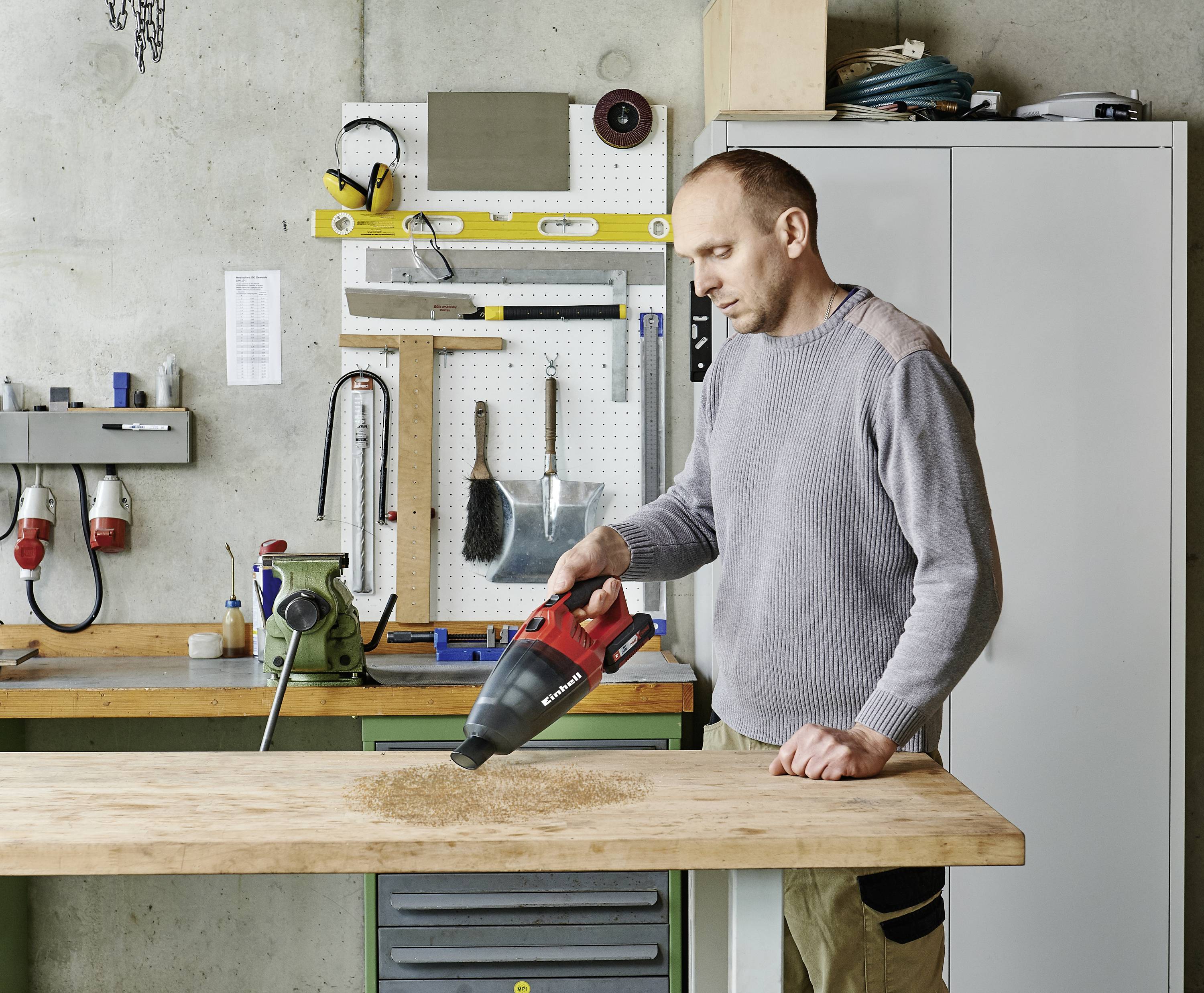 A man is using a handheld vacuum cleaner to remove wood shavings from a table in a workshop. Tools are hanging on the wall.