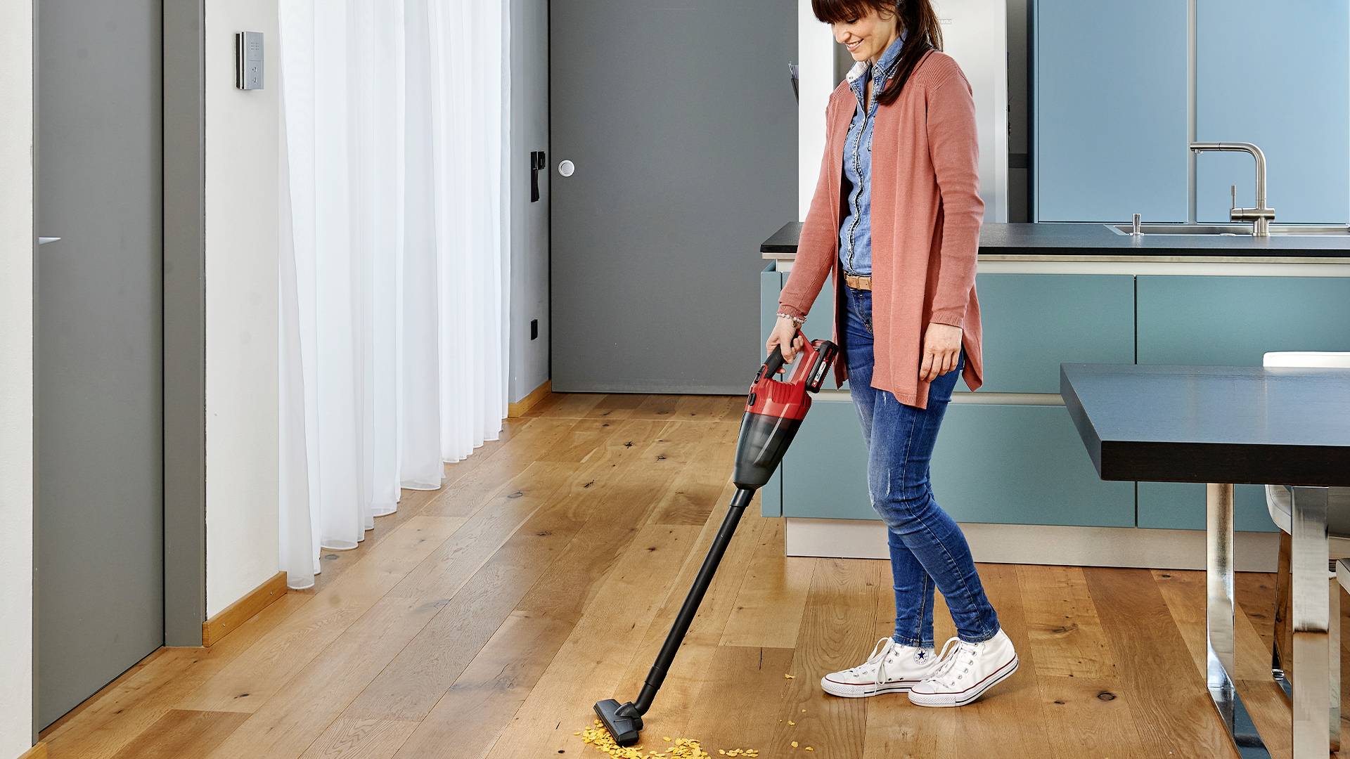A woman is cleaning crumbs from the wooden floor with a cordless vacuum cleaner in a modern kitchen.