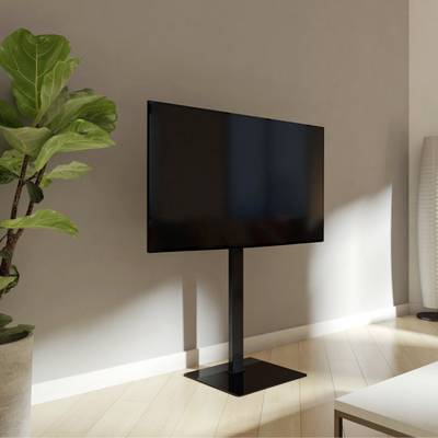 A modern television sits on a stand in a bright room, surrounded by a green plant and a stylish lampshade.