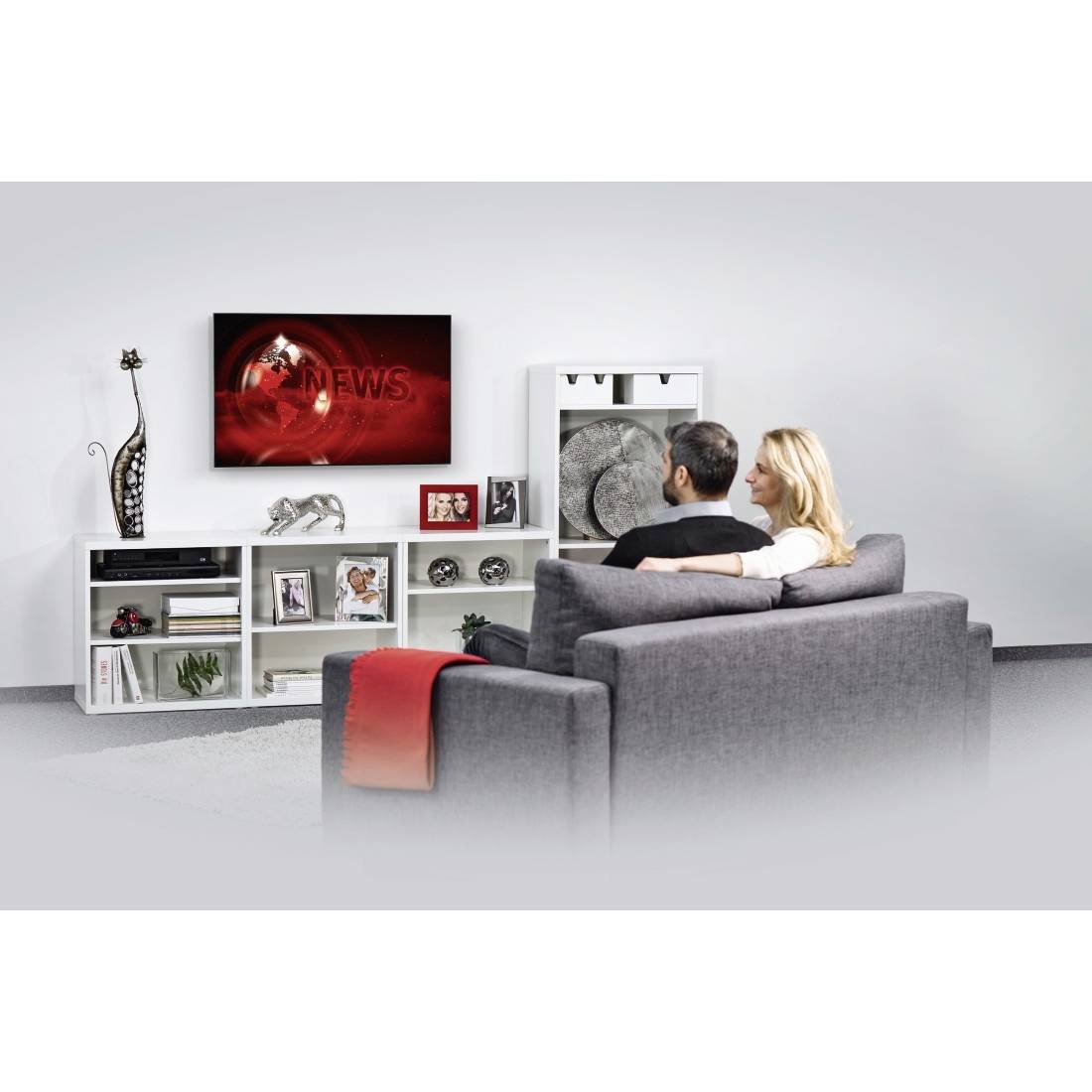 A couple sits on a sofa watching 'NEWS' on a wall-mounted flatscreen television. The room is styled in a modern interior design.