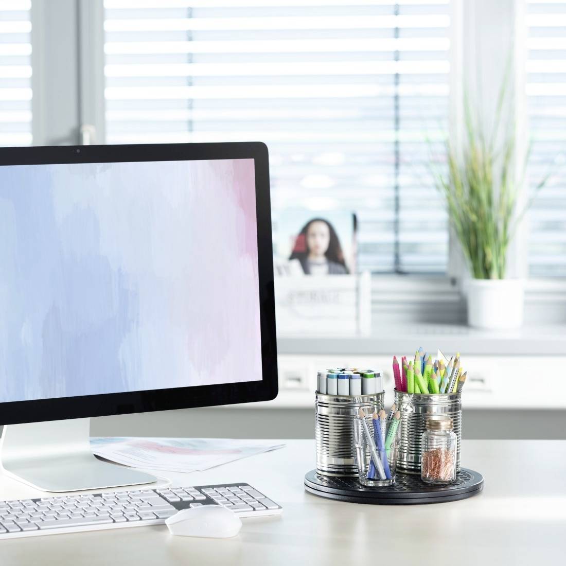A desk with a computer screen displaying pastel colours. Next to it, pens are placed in tins and plants sit on the windowsill.