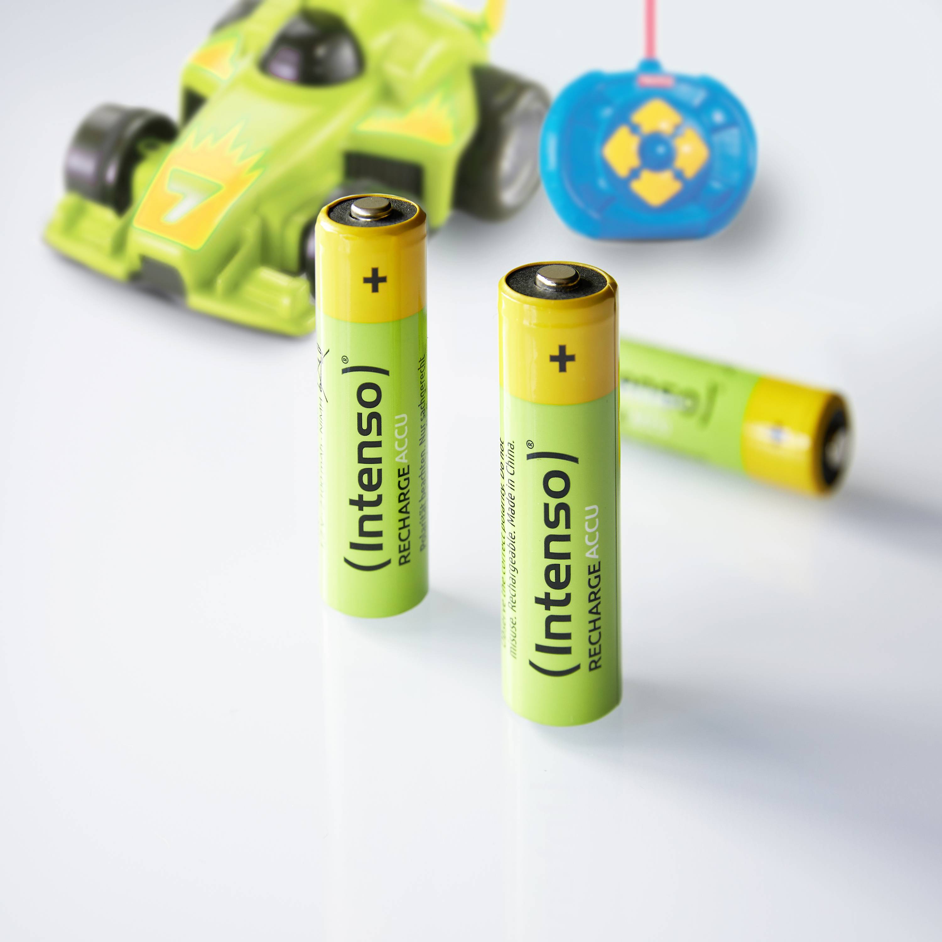 Two yellow 'Intenso' AA batteries stand upright in front of a remote-controlled toy car and a blue controller on a white background.