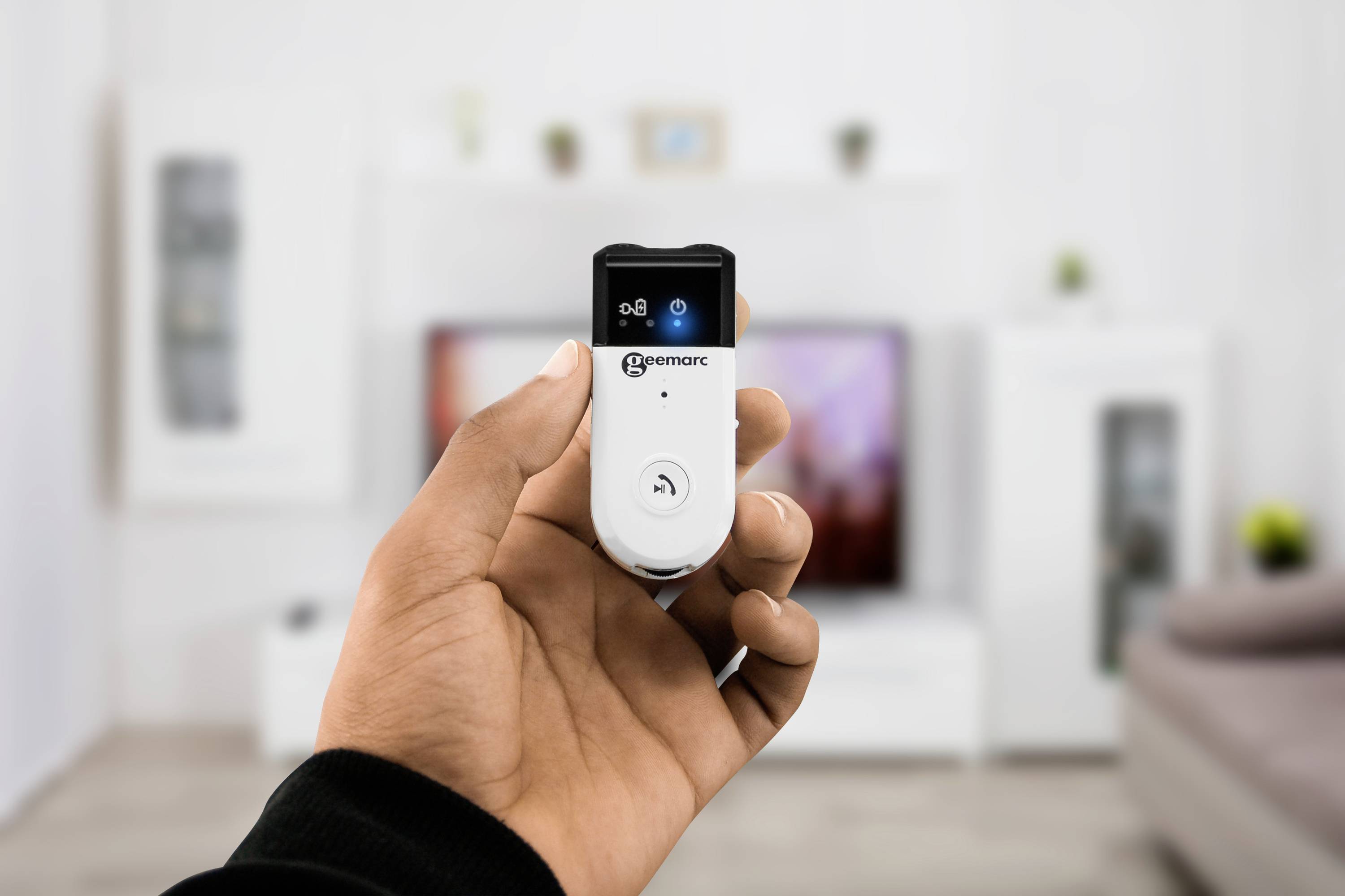 A hand is holding a small, white remote control in a living room. A television and shelves are blurred in the background.