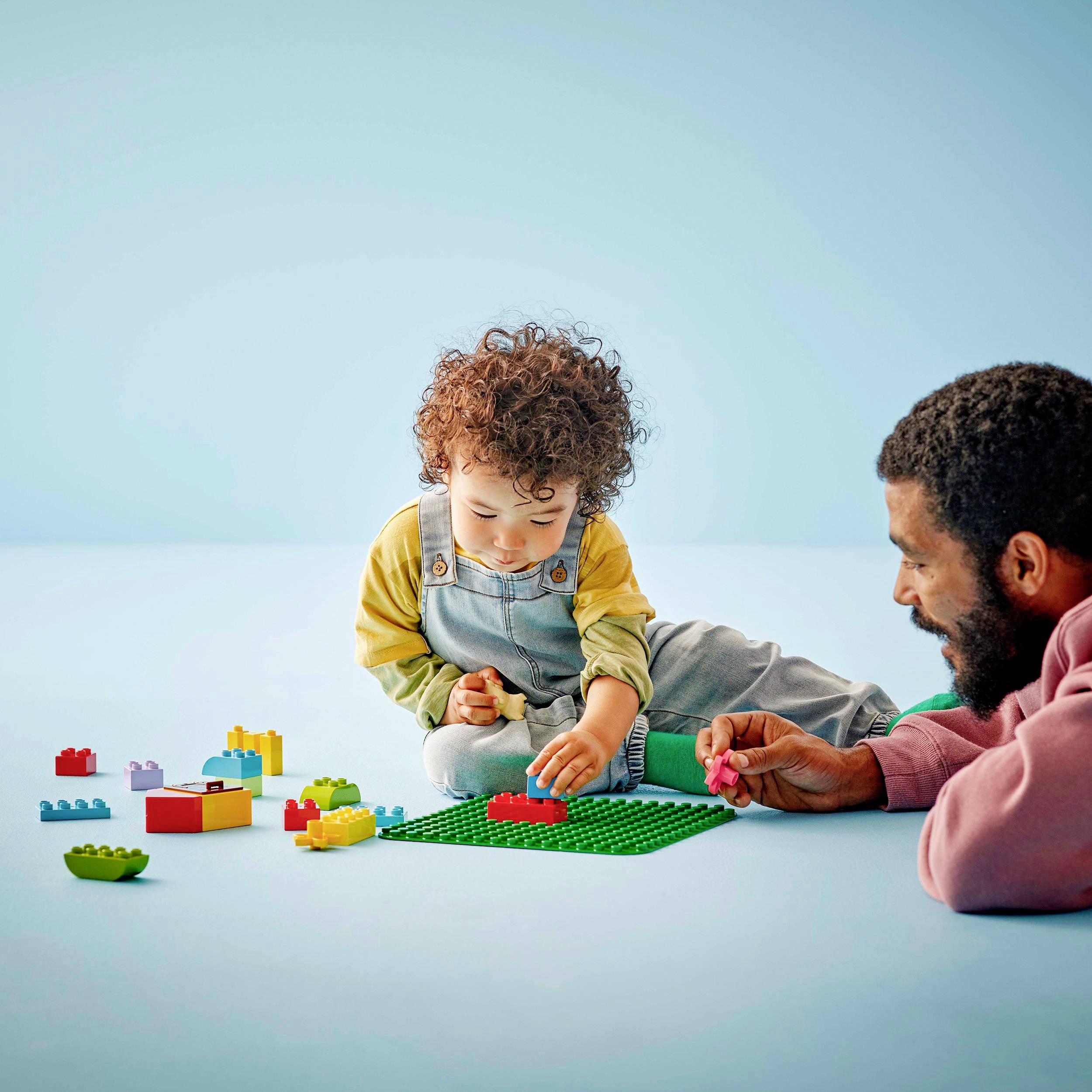 A small child is playing on the floor with coloured building blocks, while an adult watches and smiles.