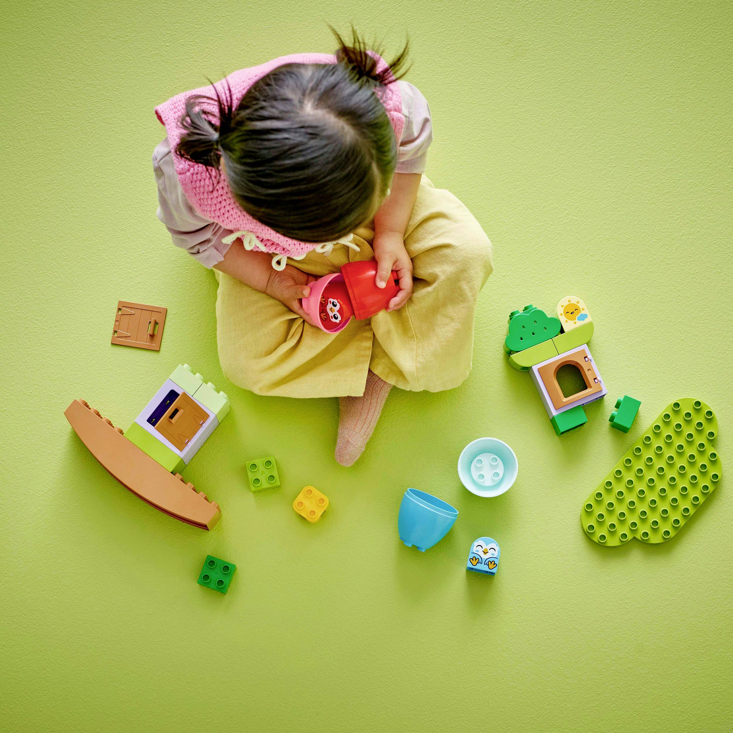 A toddler is playing on a green carpet with colourful building blocks and plastic figures, happily engaged in a creative environment.