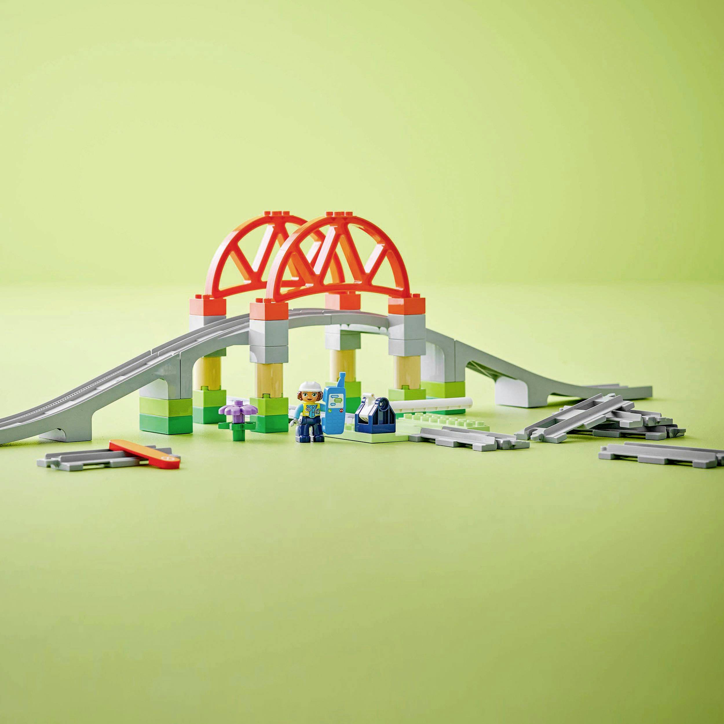 A constructed toy bridge with a red arch and grey ramps. Small figures and accessory parts are scattered on a green background.