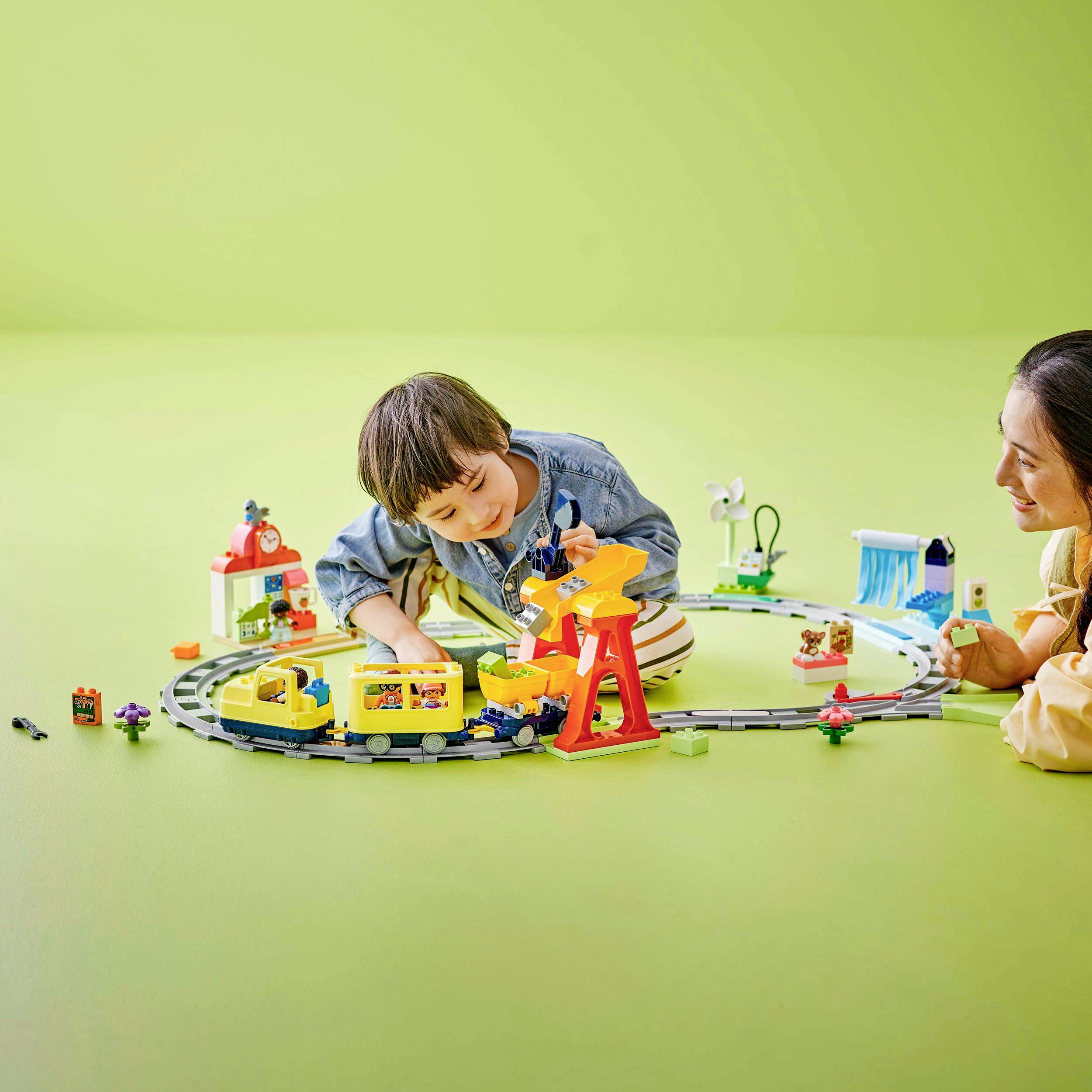 Two children are happily playing on a carpet with a toy train and figurines, surrounded by colourful toys on a green background.