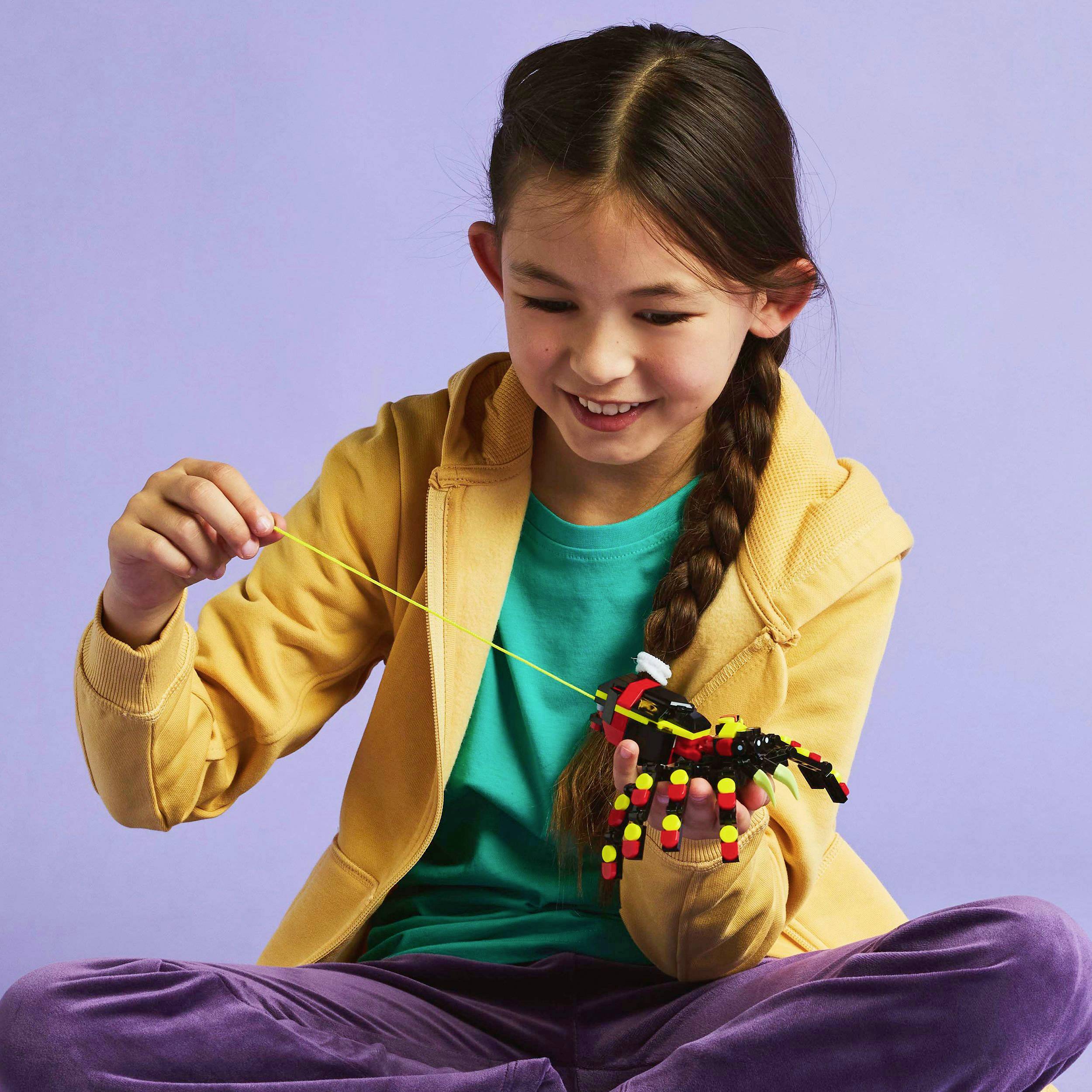 A girl in a yellow jacket is playing with a colourful robot made of building blocks, smiling against a lilac background.