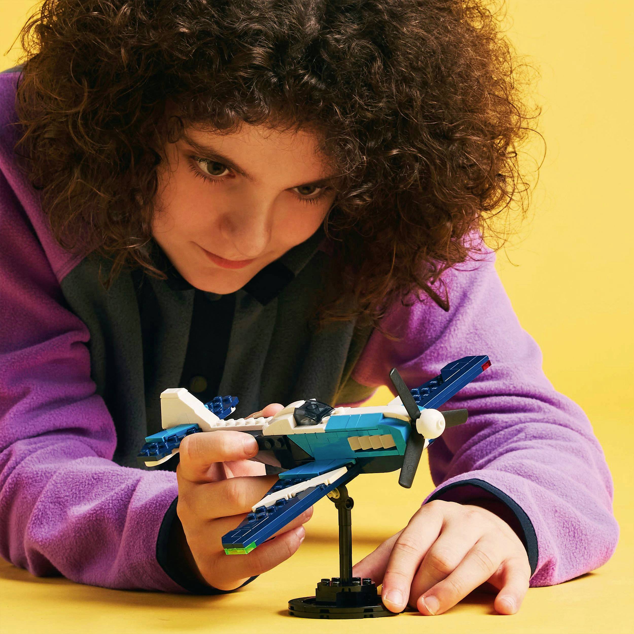 A person with curly hair is studying a small aeroplane model, composed of building blocks, on a yellow table.