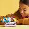 A girl smiles while looking at a small flower arrangement made of building blocks, placed on a table in a bright room.