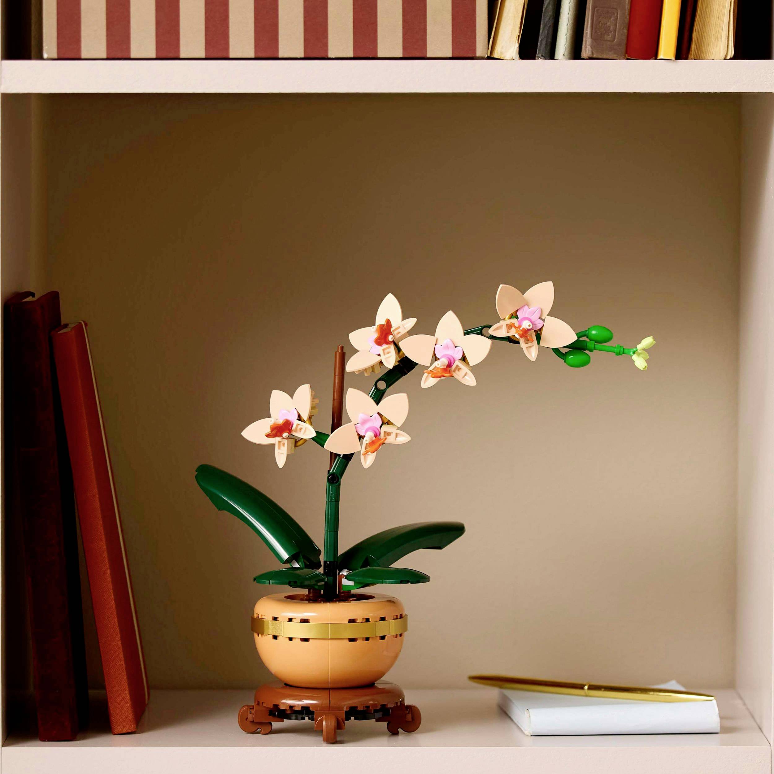 A LEGO model of an orchid in a flower pot sits on a bookshelf next to books and a closed notebook with a pen.