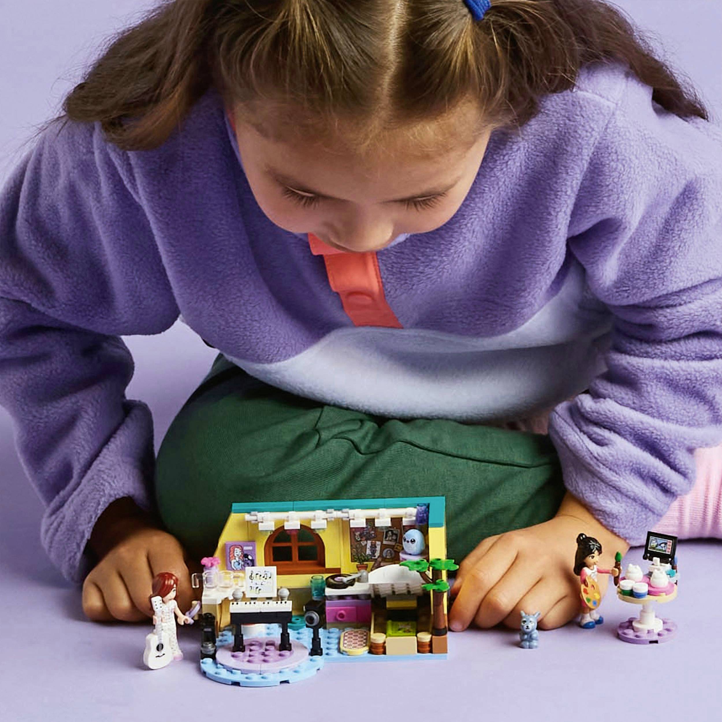 A child plays intently with a colourfully designed dolls' house made of building blocks. Small figures are positioned to the left and right.
