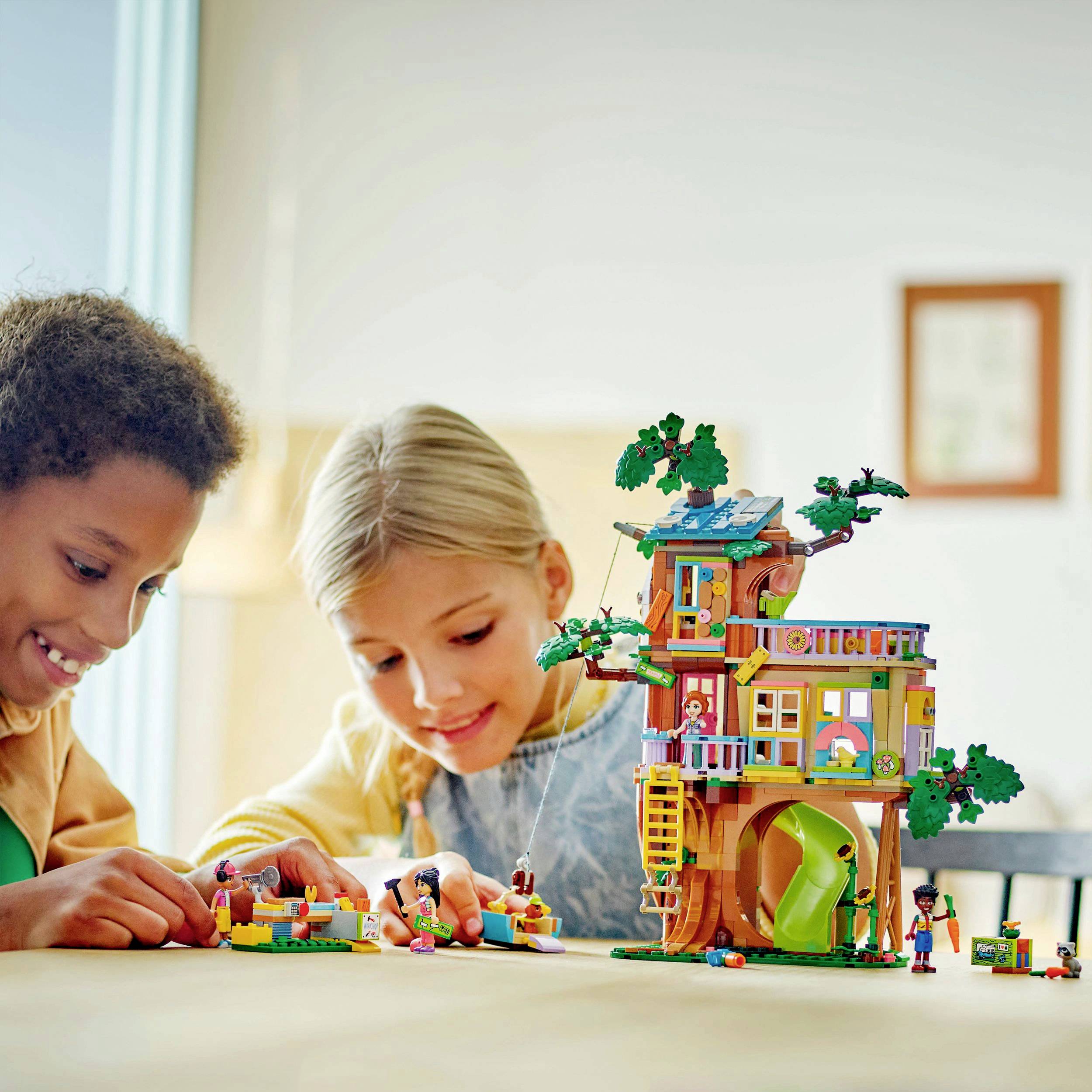 Two children are playing with a colourful Lego treehouse set on a table. The treehouse has a slide and is intricately designed.