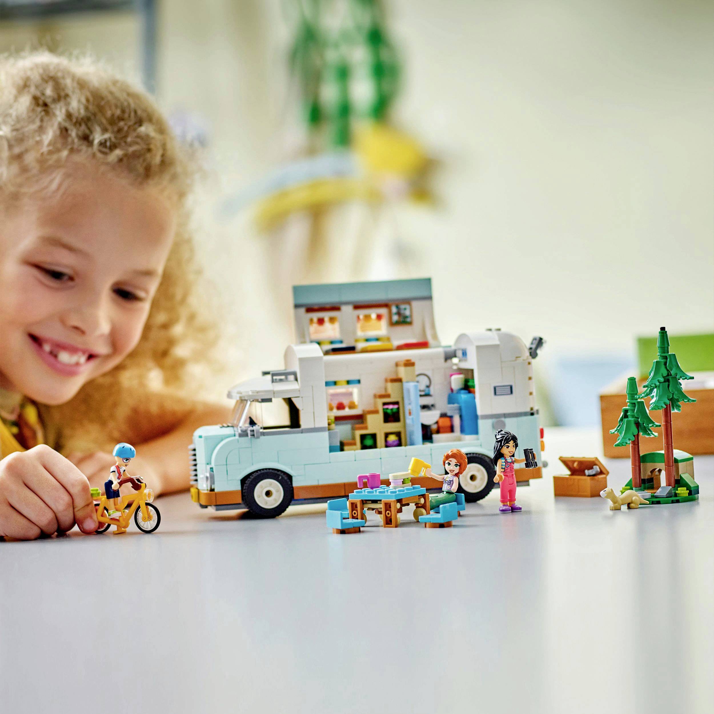 A child is playing with a model of a campervan made from building blocks, surrounded by small figures and trees on a table.