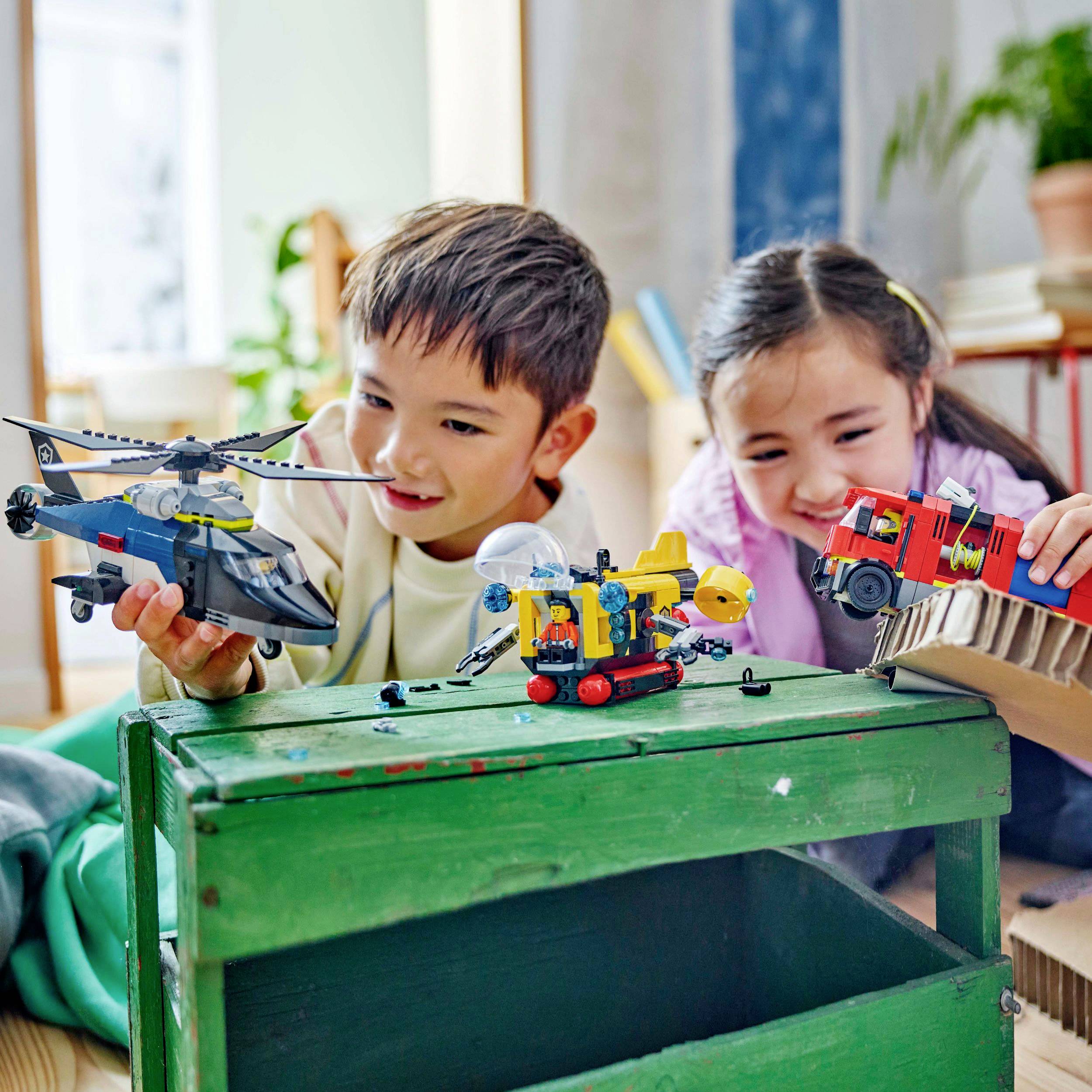 Two children are happily playing with toy vehicles on a green table in a bright room. A helicopter, a car and a lorry are visible.