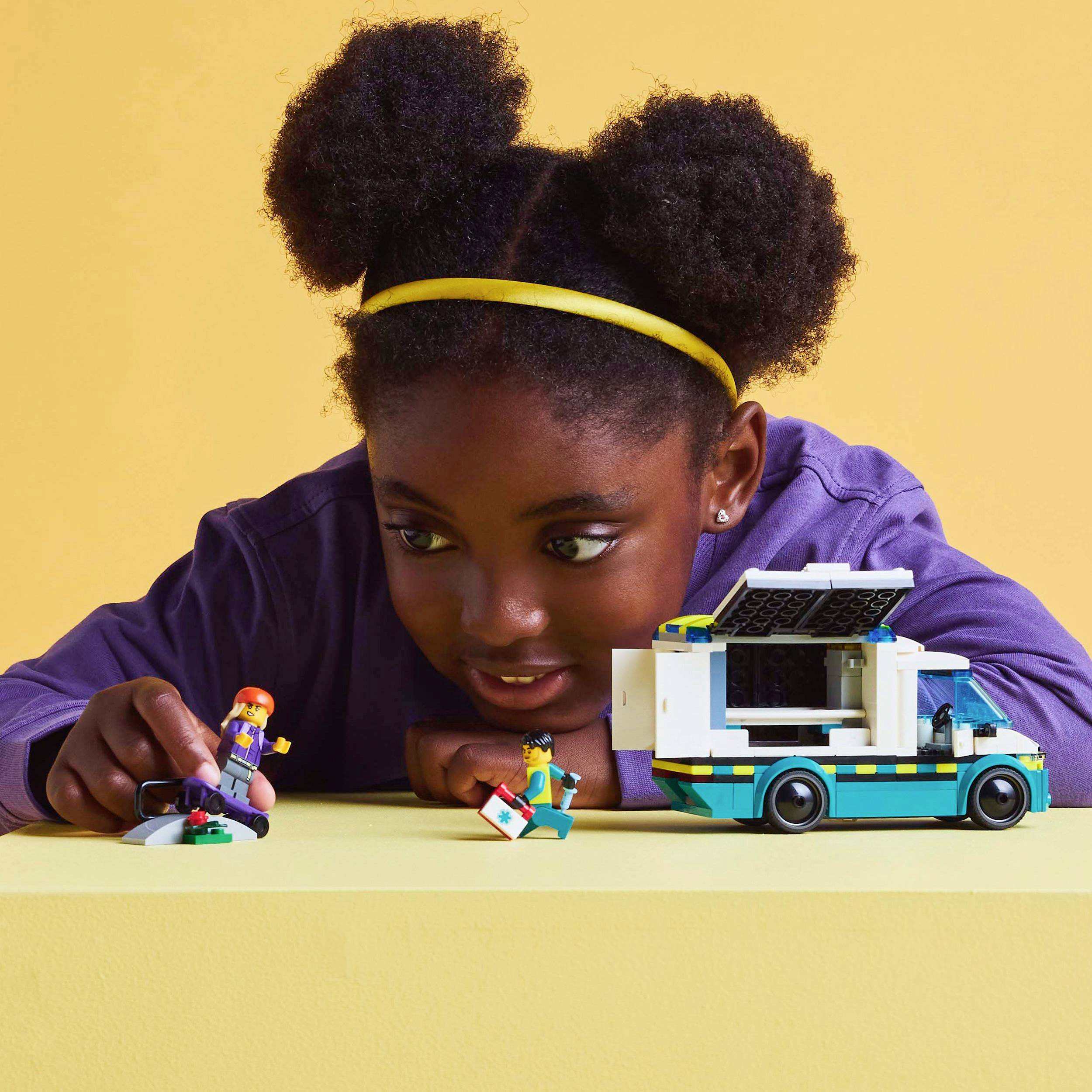 A child plays with a toy car and figurines on a table, looking at them curiously. The background is yellow-orange.