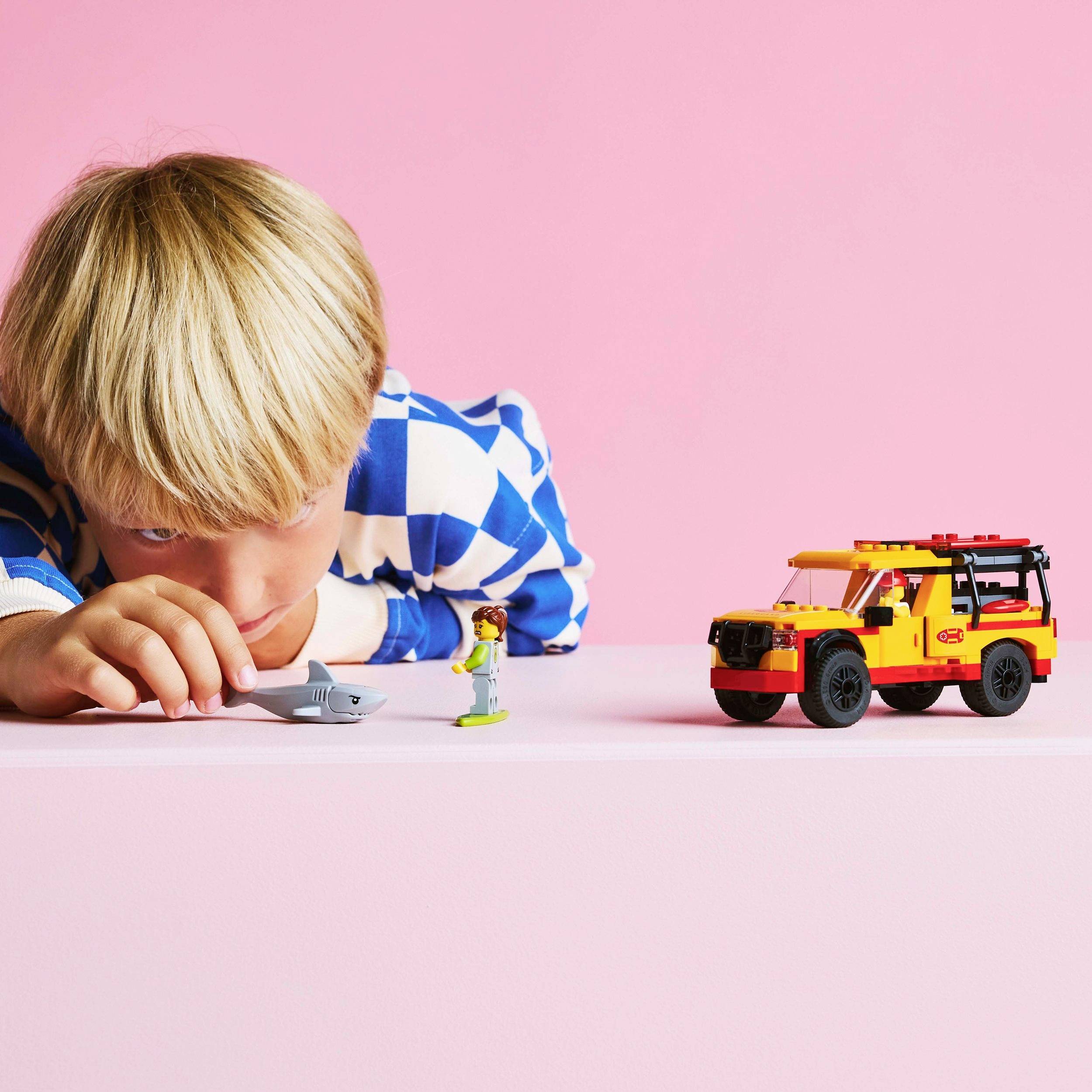 A child is playing with toy figures and a yellow toy car on a table in front of a pink background.