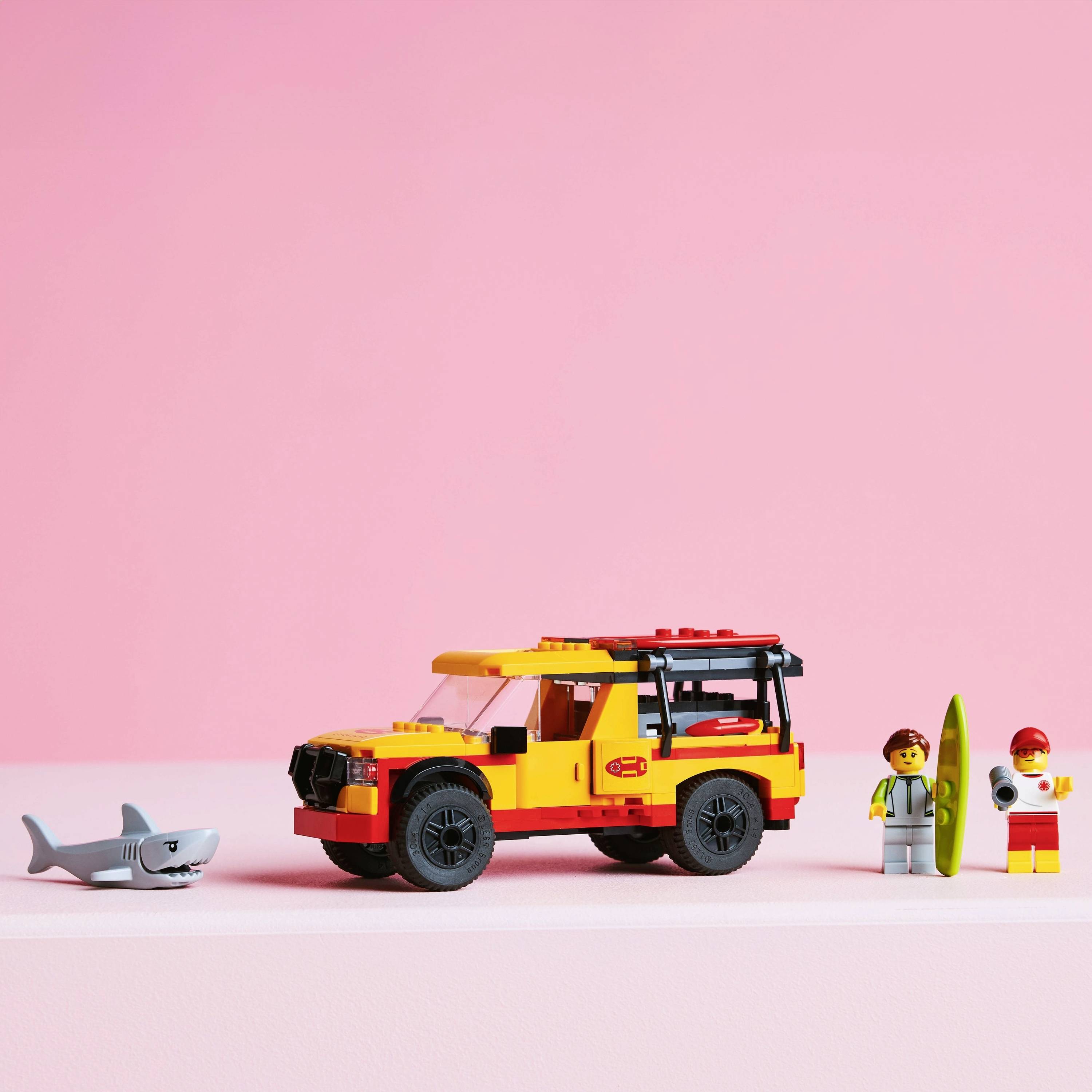 Toy ambulance with two figures and a shark on a pink background. One figure is holding a surfboard.
