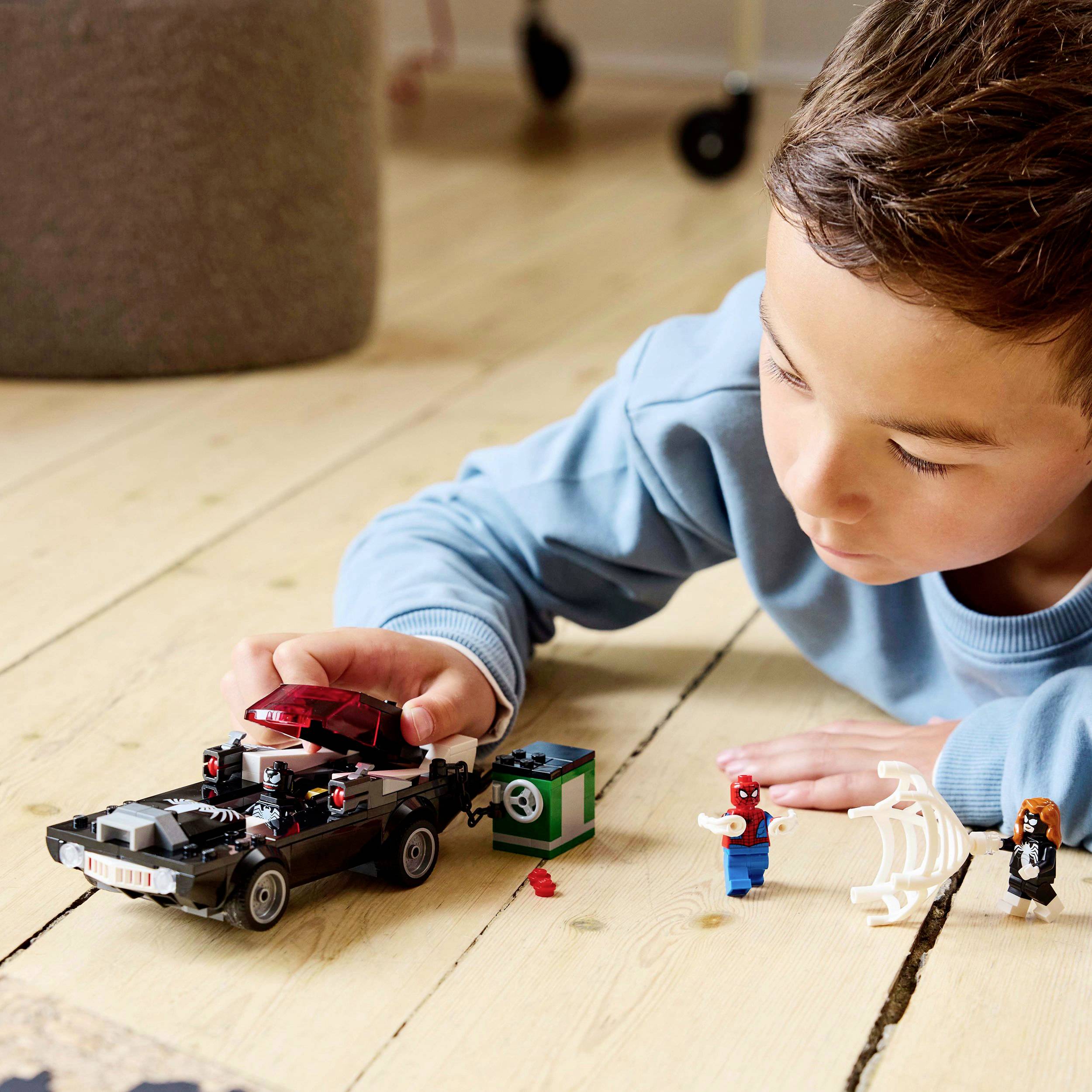 A child is playing on a wooden floor with a small car construction made of building blocks and figures, including a skeleton and a cardboard box.
