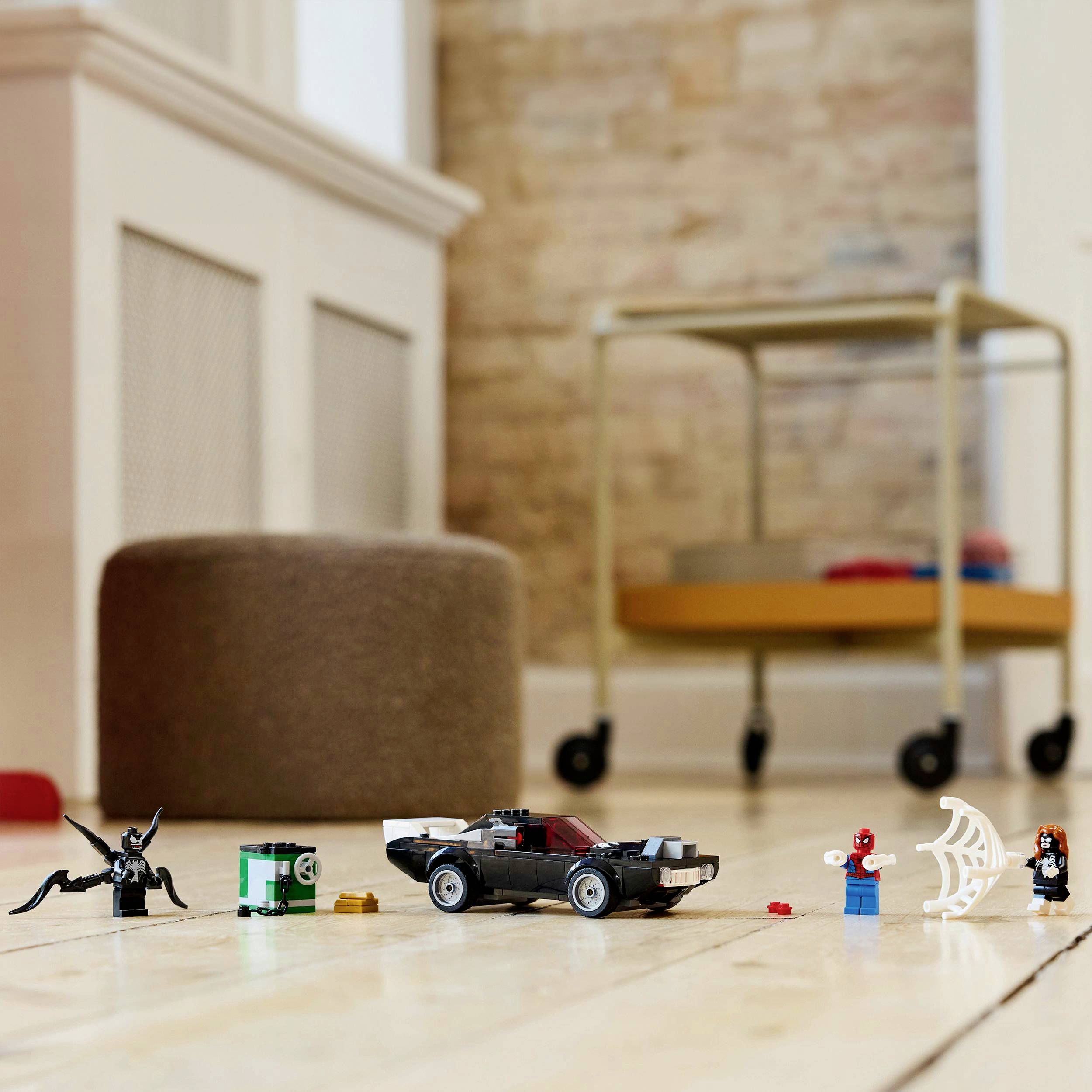 Toys on a wooden floor: A small black toy car, block-like figures, and a spider's web. Background shows furniture and a brick wall.