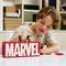 A child plays with LEGO and builds 'MARVEL' letters out of red bricks. Next to them, superhero minifigures stand on a table.