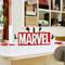 On a table sits a LEGO logo spelling out 'MARVEL'. Small LEGO figurines of superheroes are positioned on it. In the background, a shelf with boxes can be seen.