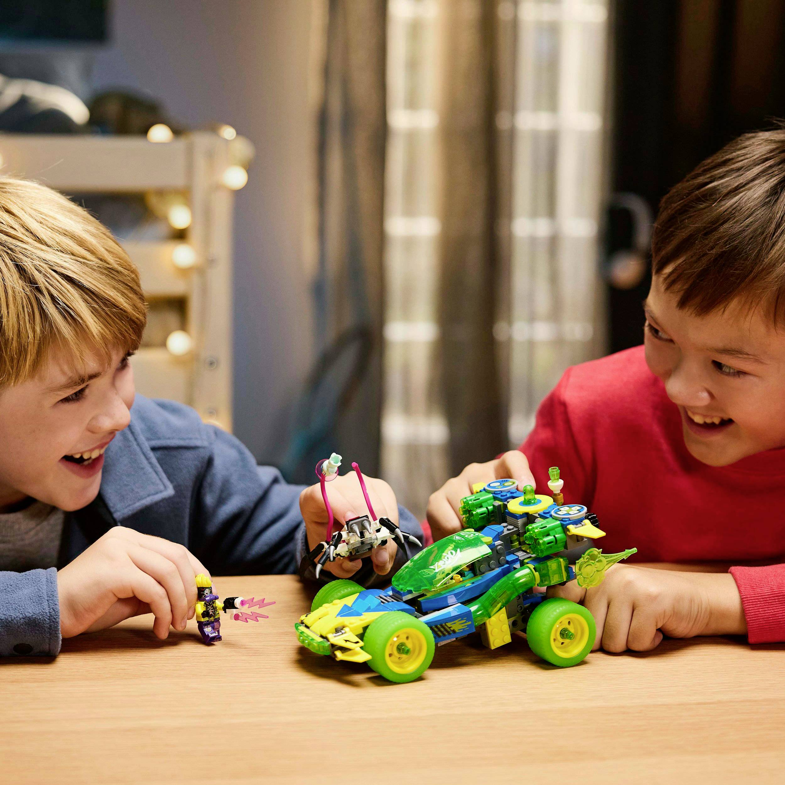 Two children are playing with a remote-controlled car on a table. The car is colourful and has large wheels. Both children are smiling.