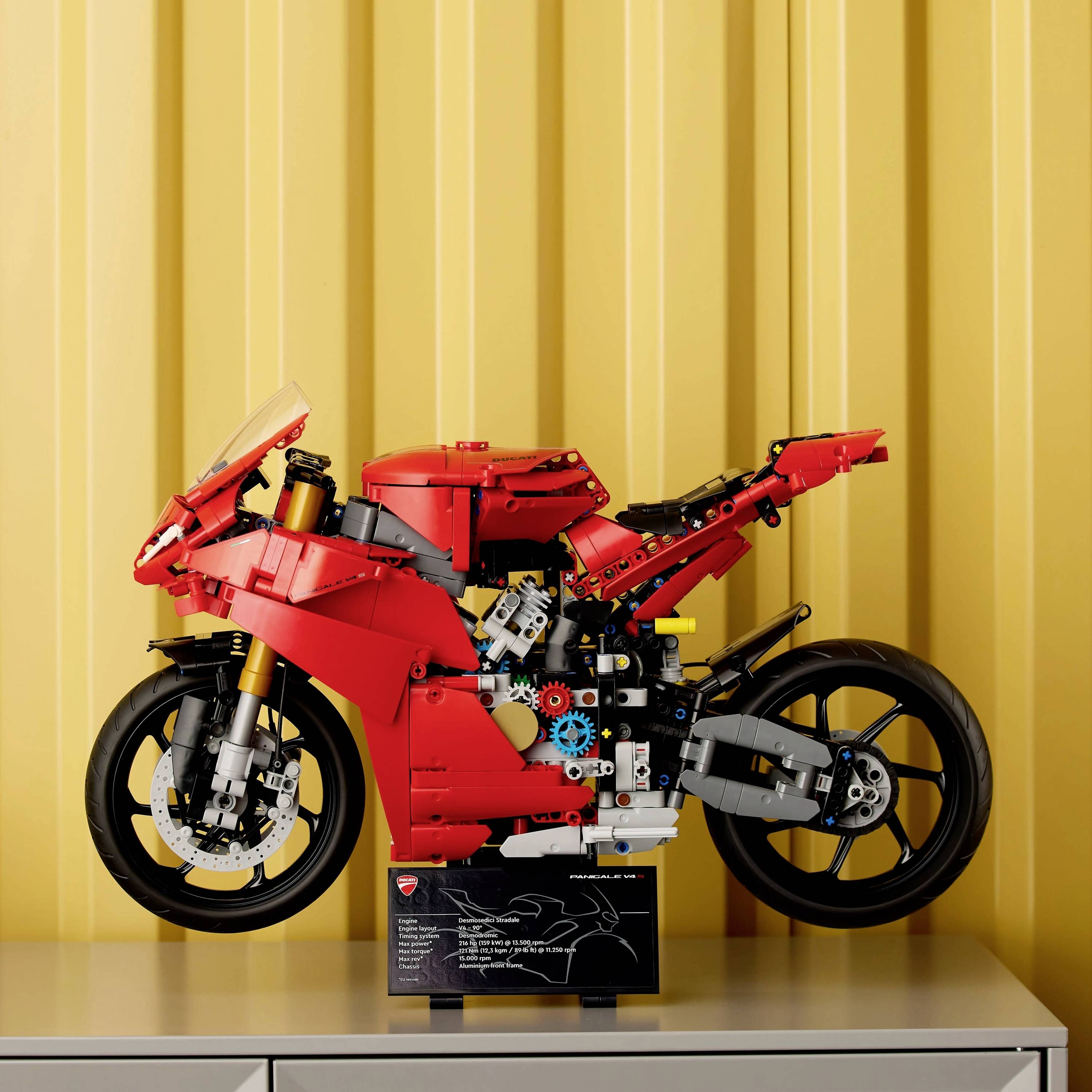 A red LEGO motorcycle model stands on a table in front of a yellow background. The model shows detailed mechanical parts.