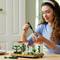 A woman is smiling as she assembles a LEGO set with a botanical and architectural theme at the table.
