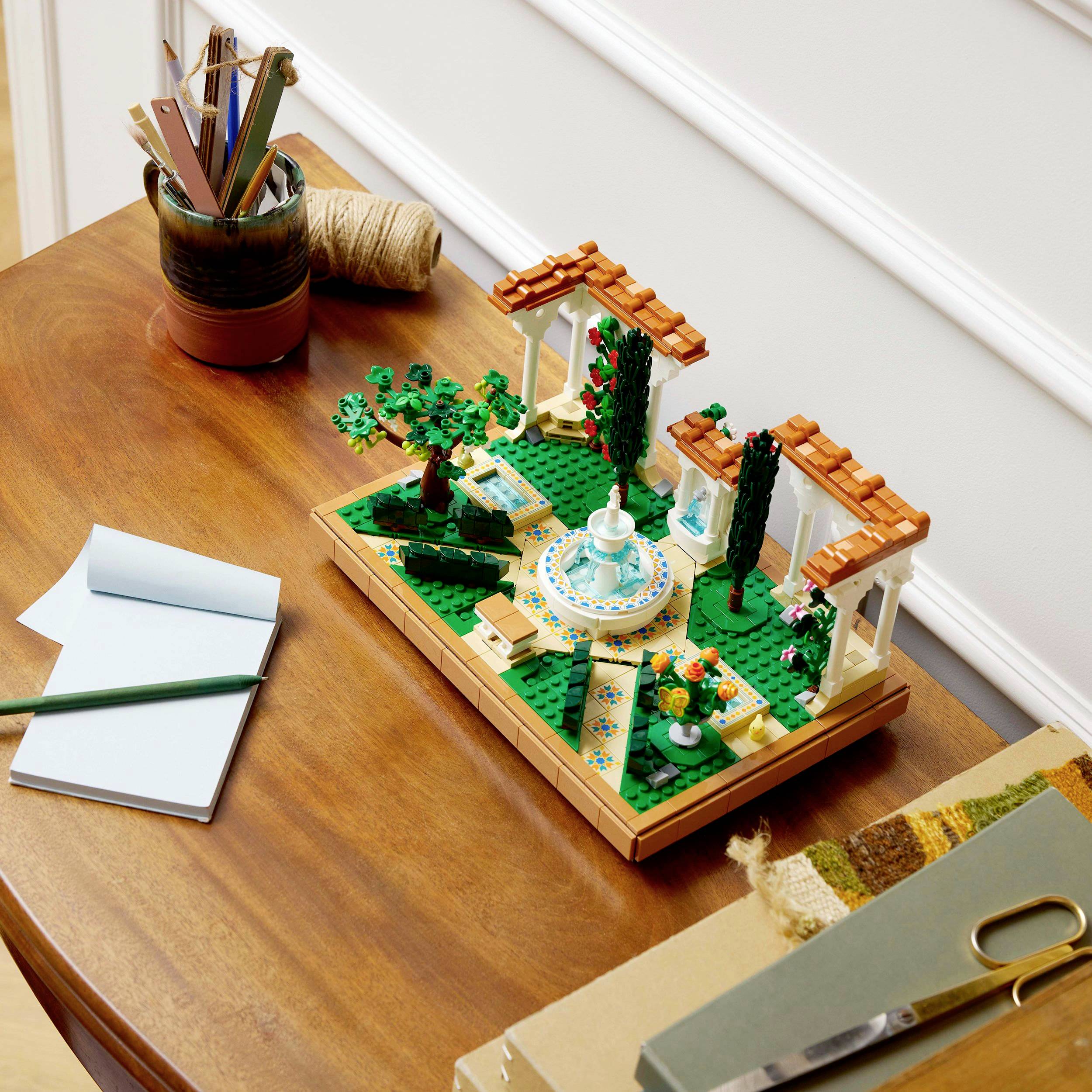 Lego set on a table, designed as a Mediterranean garden with trees, a terrace, and a small fountain. Notebook and pens in the background.