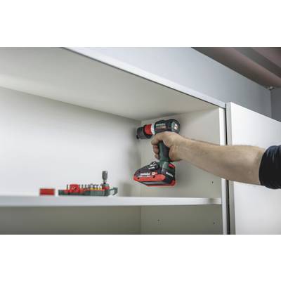 A person is holding a cordless drill and assembling a shelf. Screws and drill bits are in the foreground.