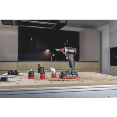 A cordless drill sits on a table with several attachments. In the blurred background, a modern kitchen is visible.