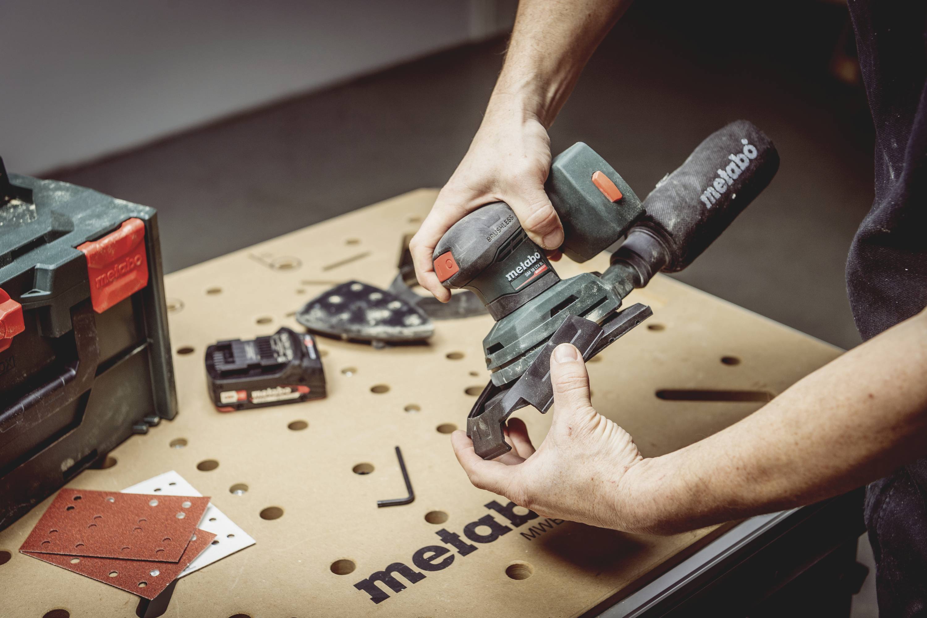 A person is operating an electric grinding machine on a workbench. Surrounded by sandpaper and a toolbox.