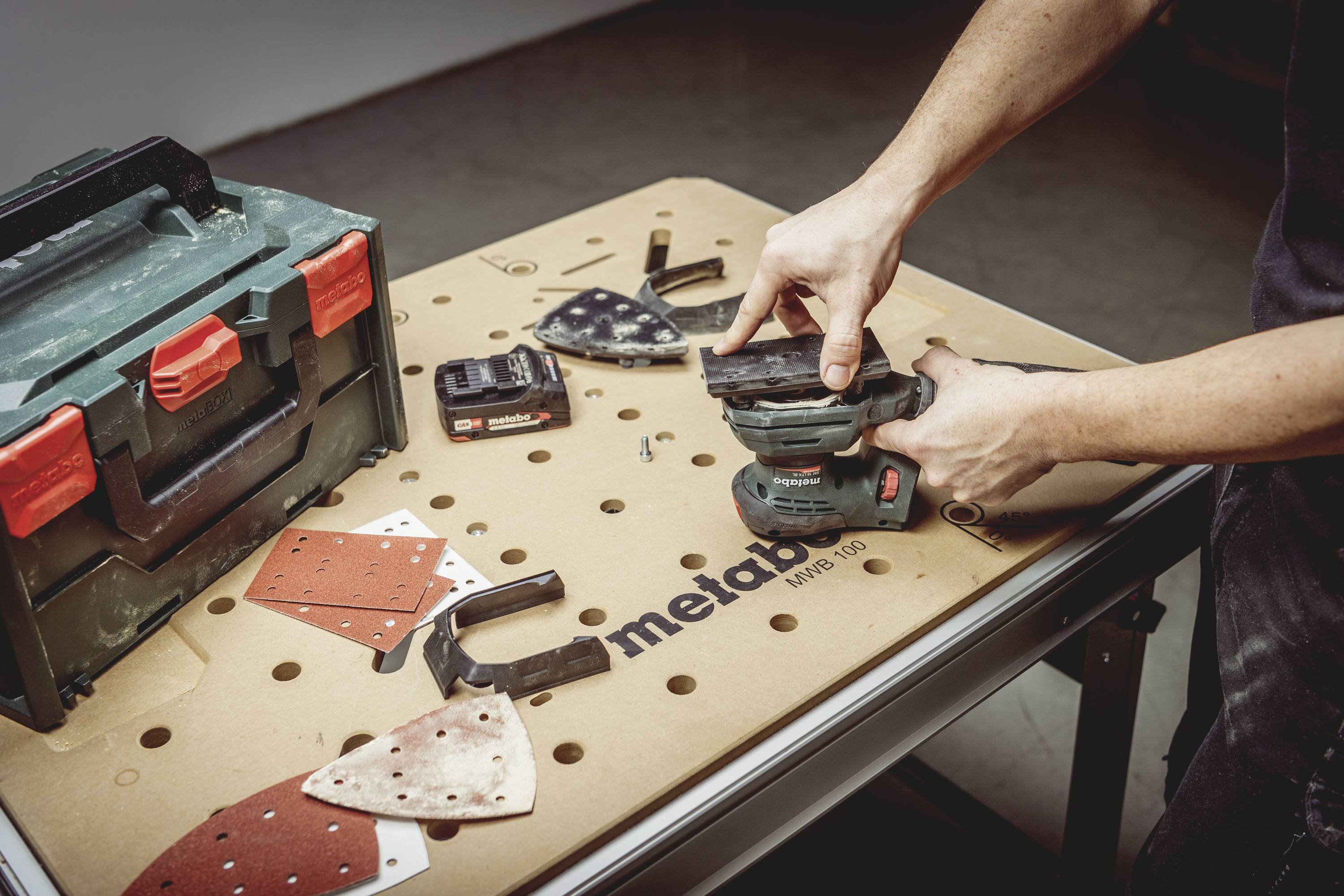 A person is using a grinding tool at a table with several sanding pad attachments and a toolbox.