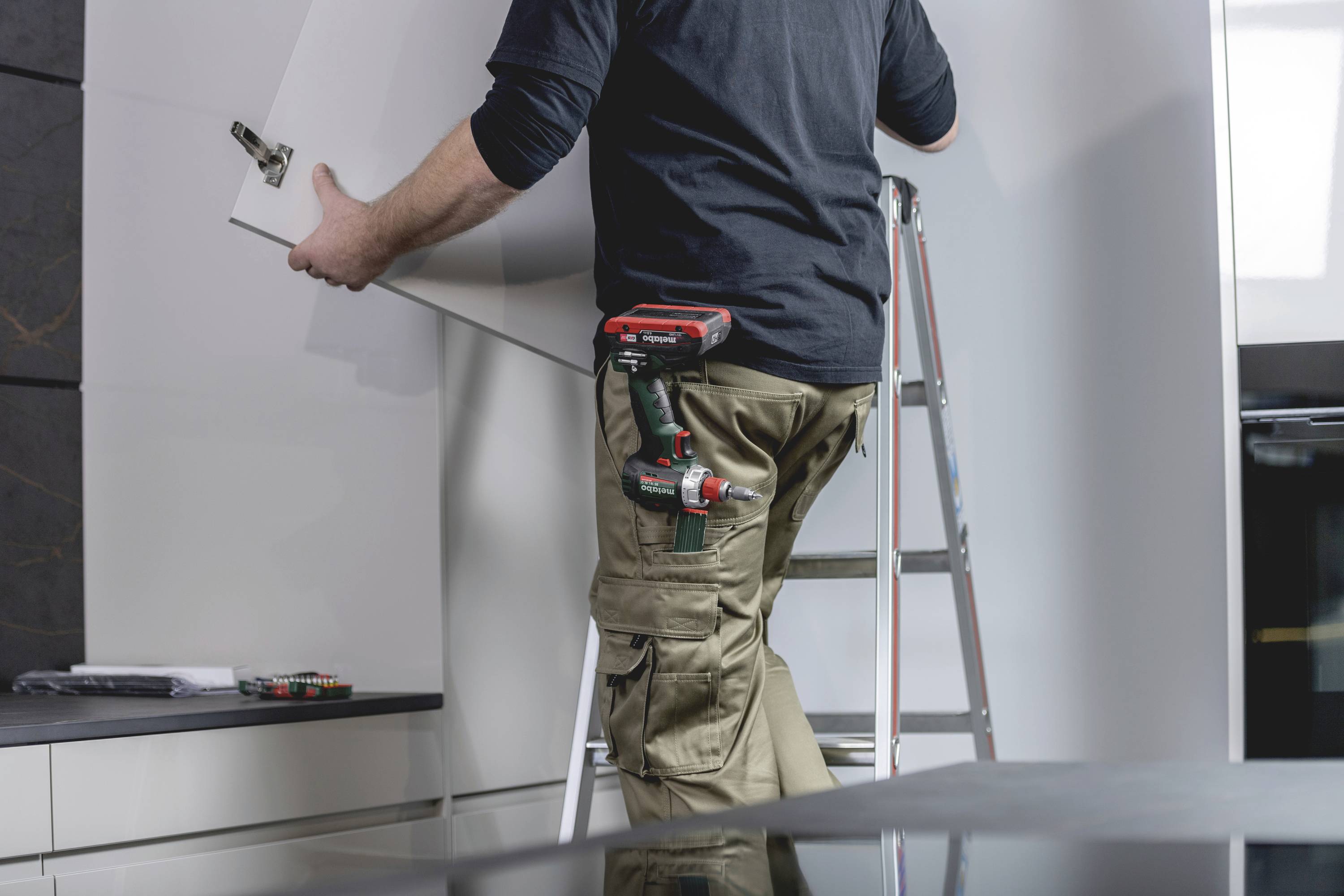 A tradesman is standing on a ladder and installing a cabinet door in a modern kitchen. A cordless drill hangs from his trouser pocket.