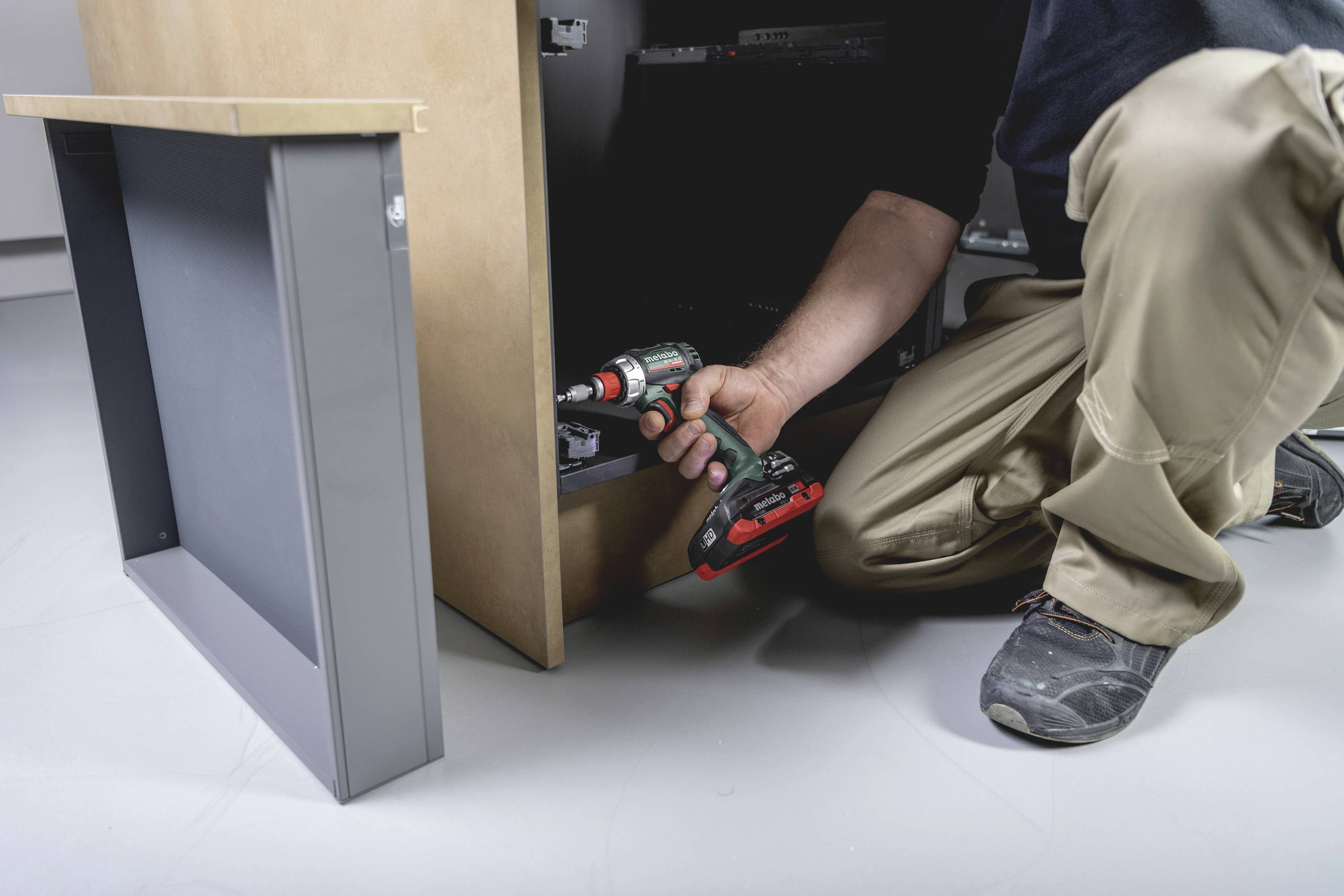 A person is kneeling on the floor and using a black and red cordless drill to assemble a piece of furniture or a wardrobe.