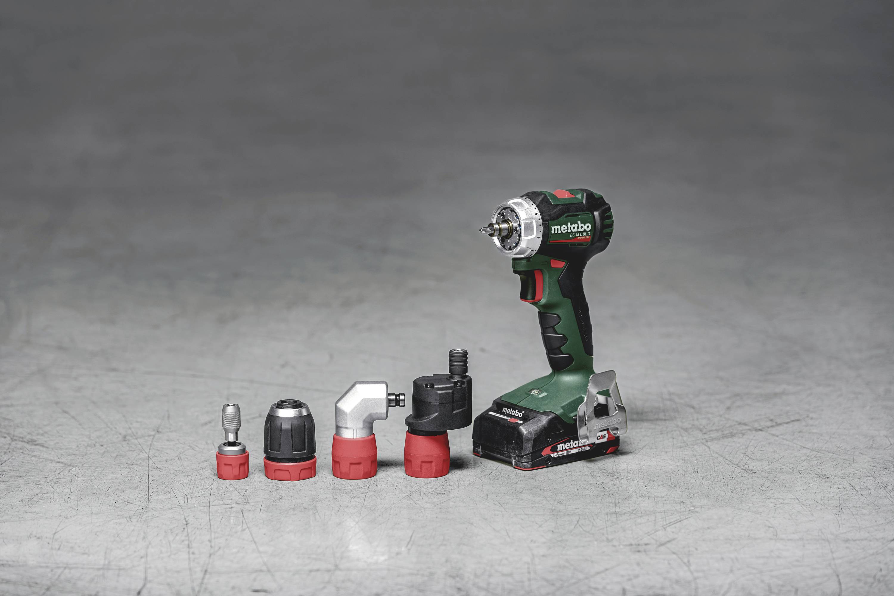 A green cordless drill with four different attachments on a grey surface.