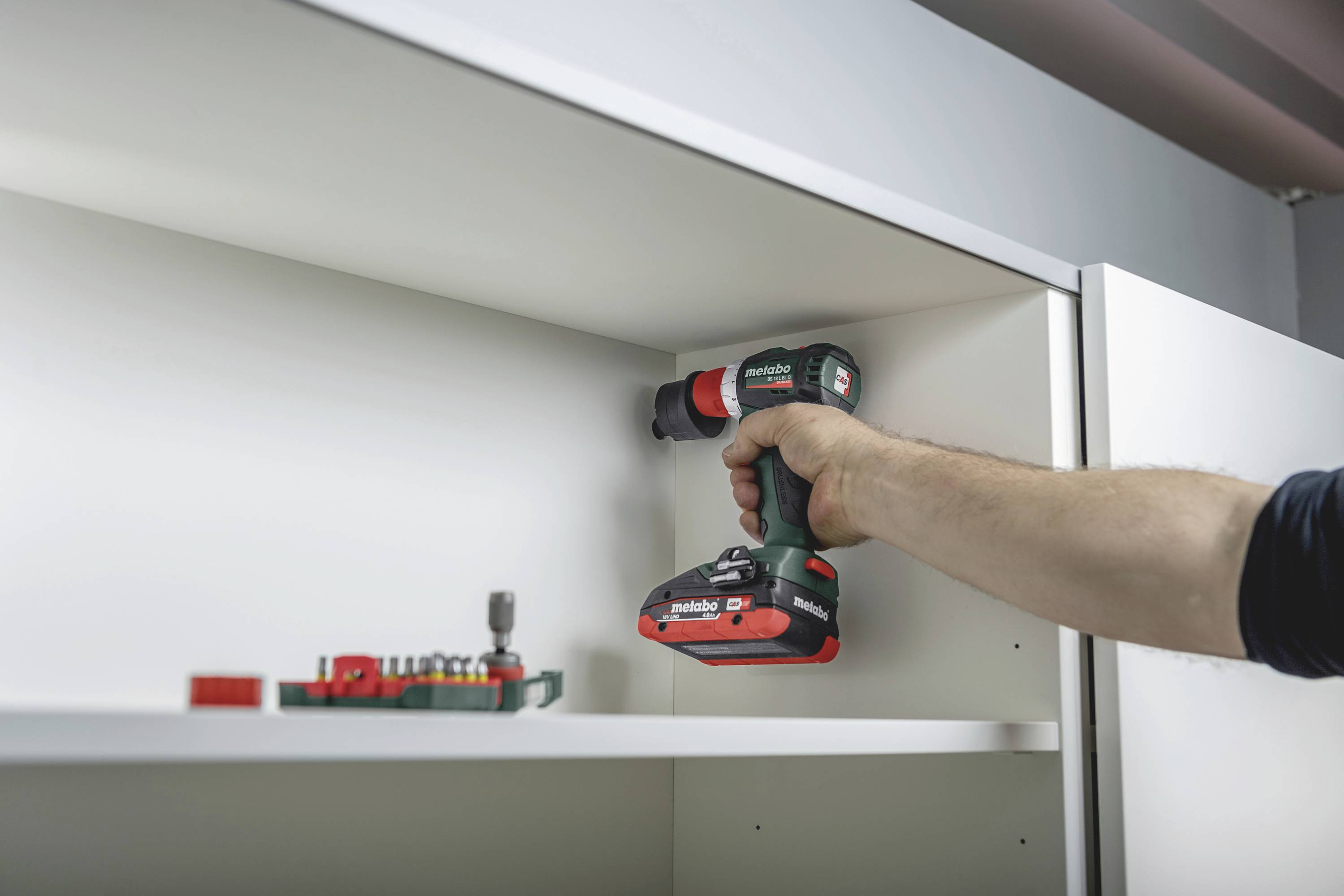 A hand is using a cordless drill to assemble a shelf. Screws and drill bits are visible in the foreground.