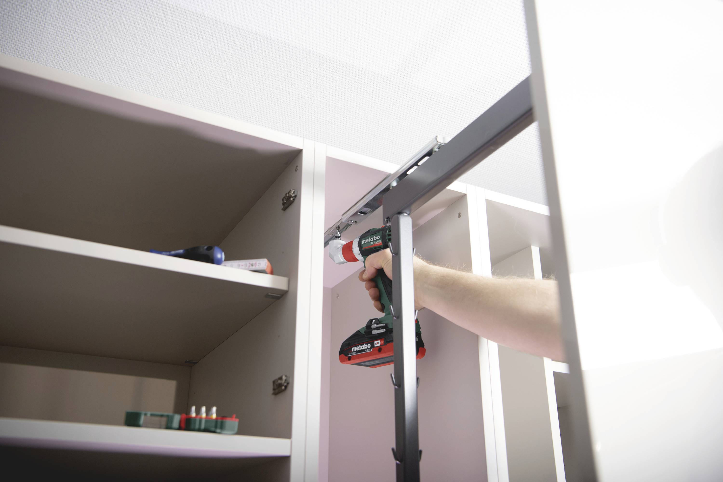 A person is using a cordless drill to mount a shelf on a light-coloured wall. Tools are lying on the shelf.